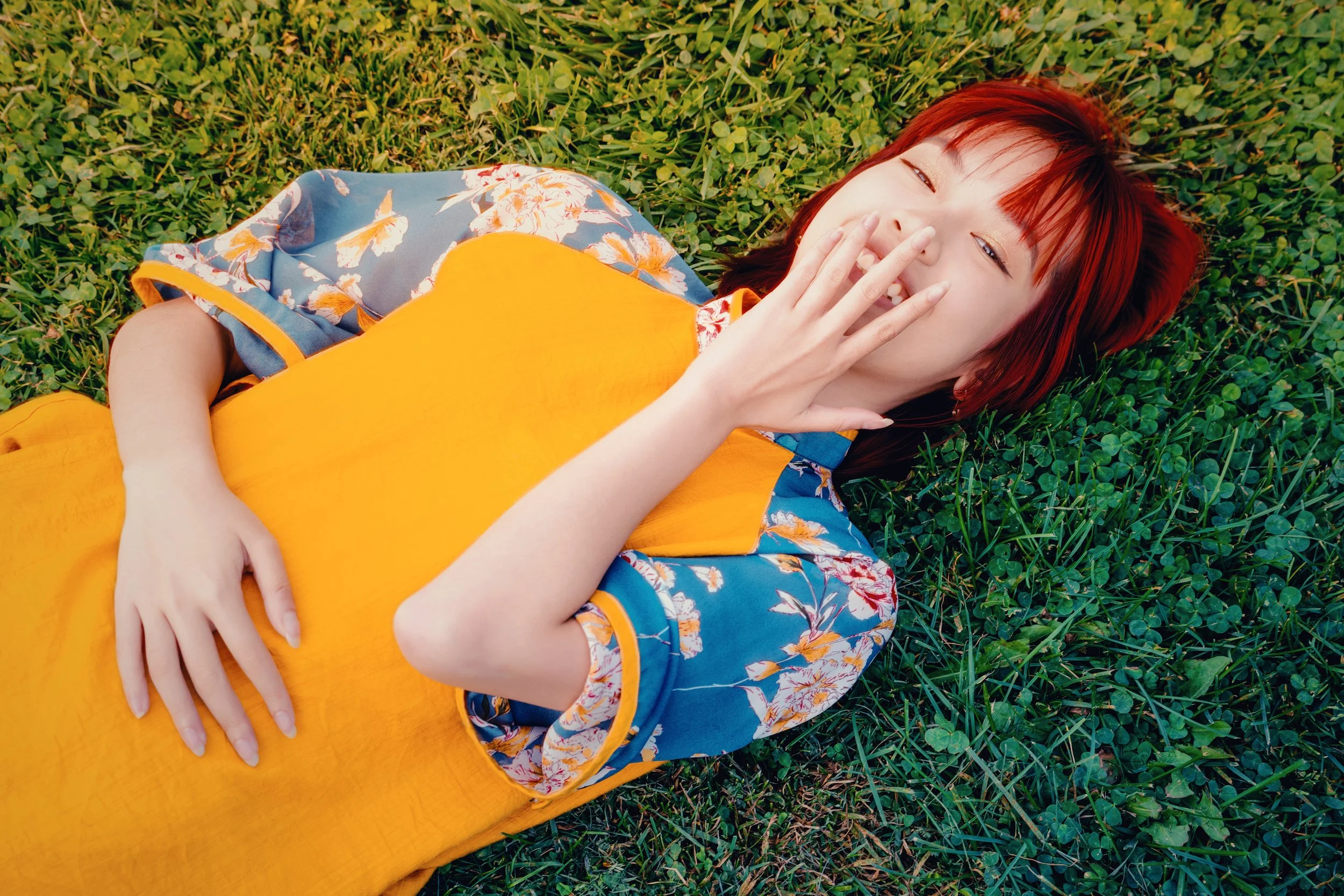 A person with red hair lying on the grass, covering their mouth with one hand, wearing a colorful blue floral shirt and an orange dress.