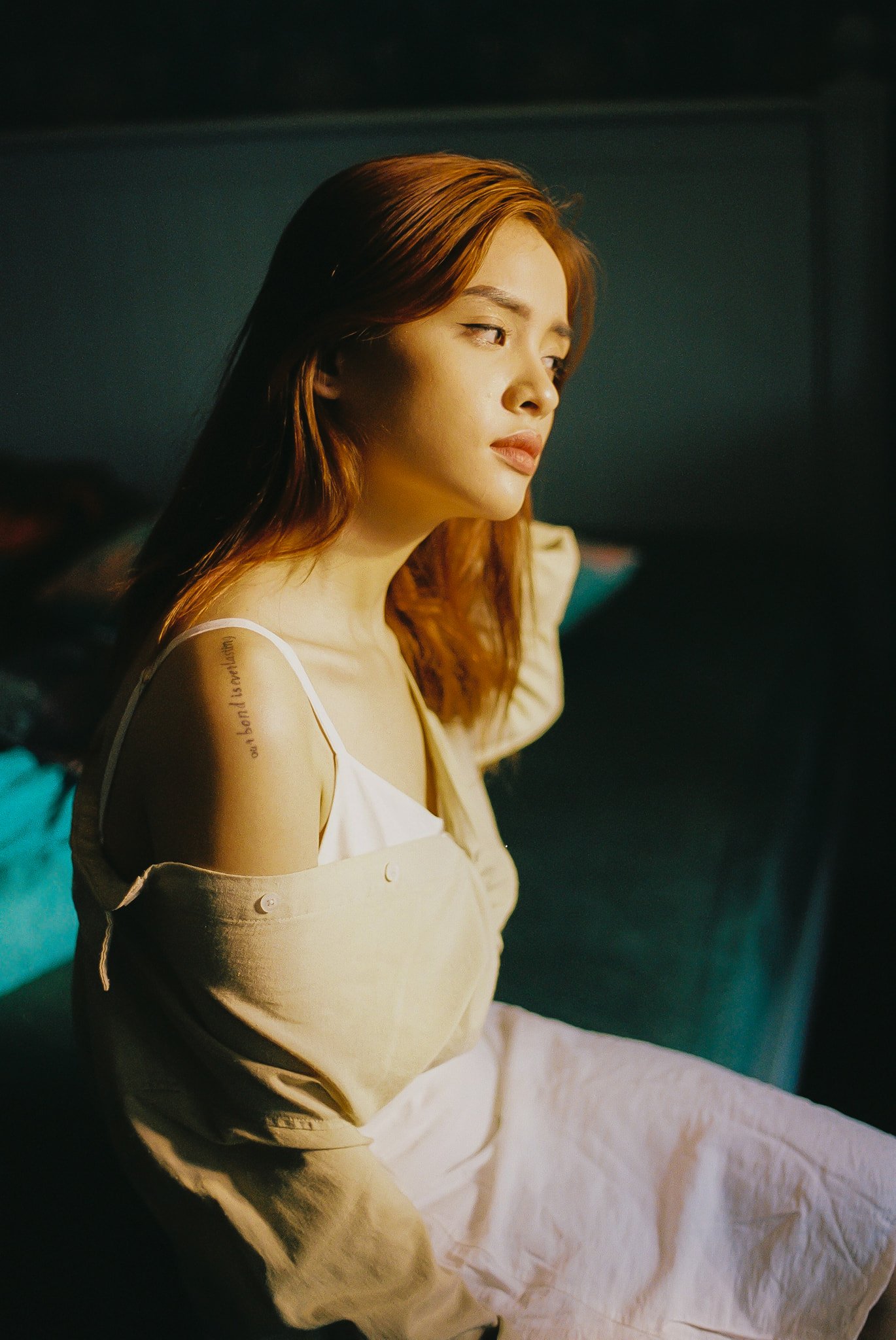 A woman with reddish hair sitting on a bed, looking away thoughtfully. She is wearing a beige jacket off her shoulders, revealing a white top underneath.