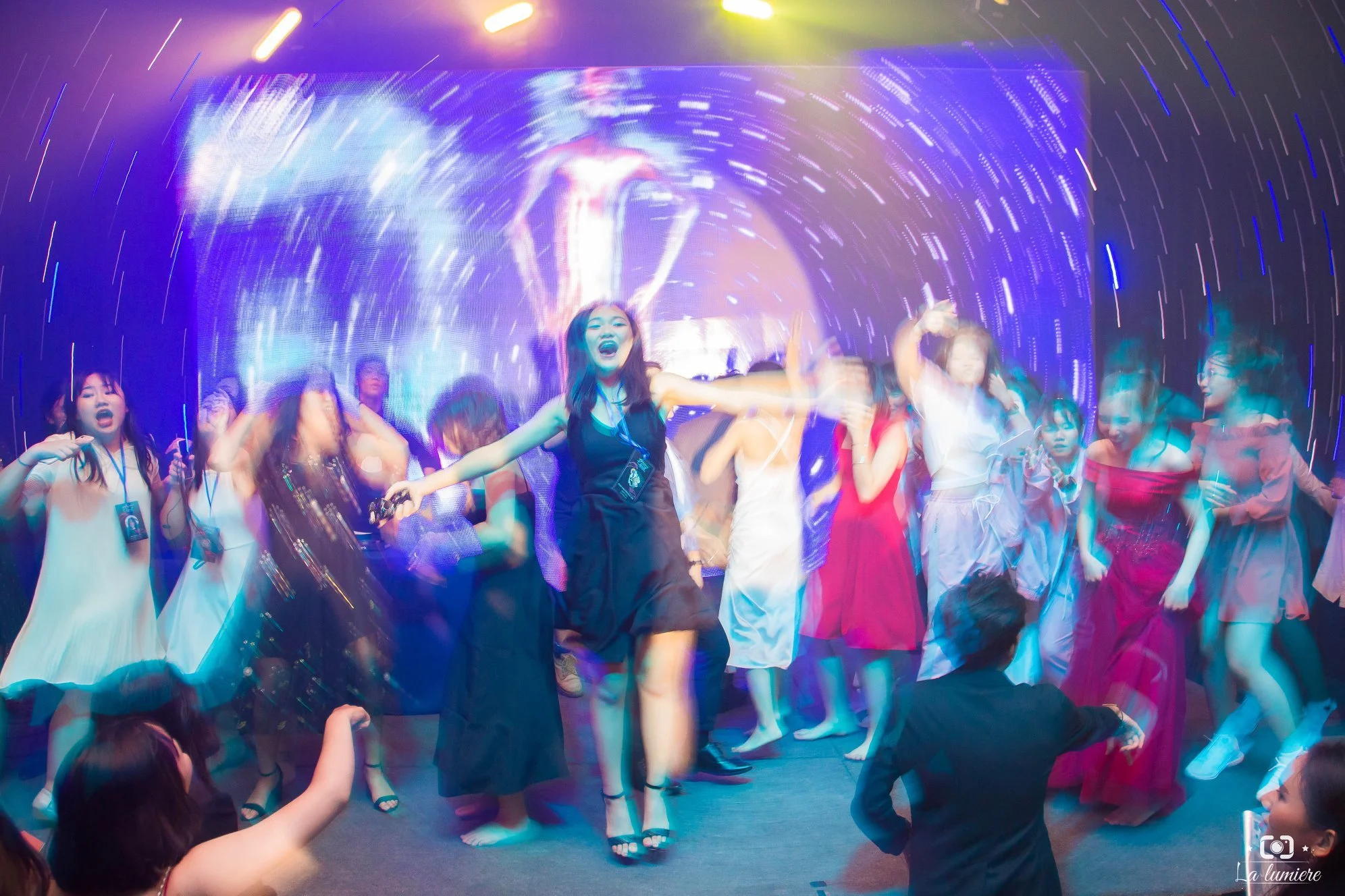 Group of young women dancing and celebrating at a party with colorful lights and a digital display in the background.