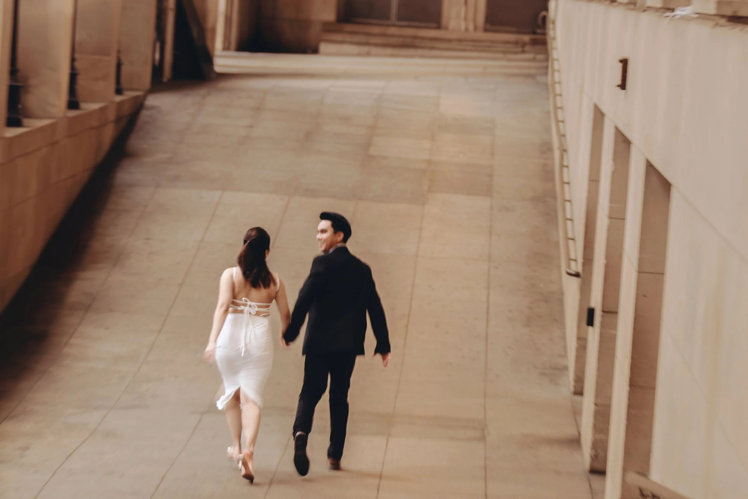 A man and woman holding hands and walking up a wide indoor staircase, the woman in a white dress and the man in a dark suit, with a beige stone wall on the right and wooden panels on the left.
