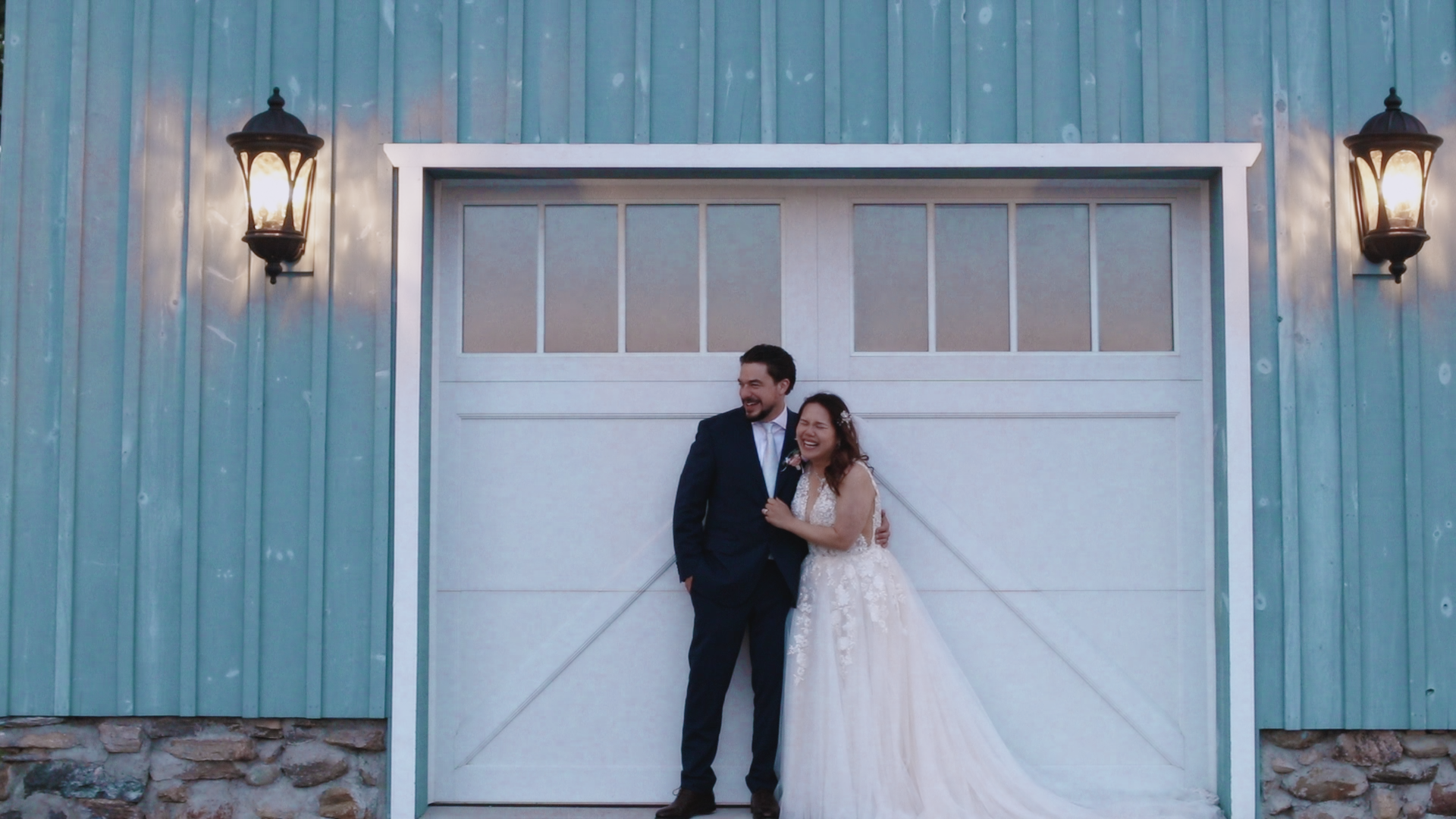 A newlywed couple standing in front of a white garage door, smiling and embracing. The groom is wearing a navy blue suit, and the bride is dressed in a lace wedding gown. The scene is lit by two black lantern-style outdoor wall lights on a blue metal