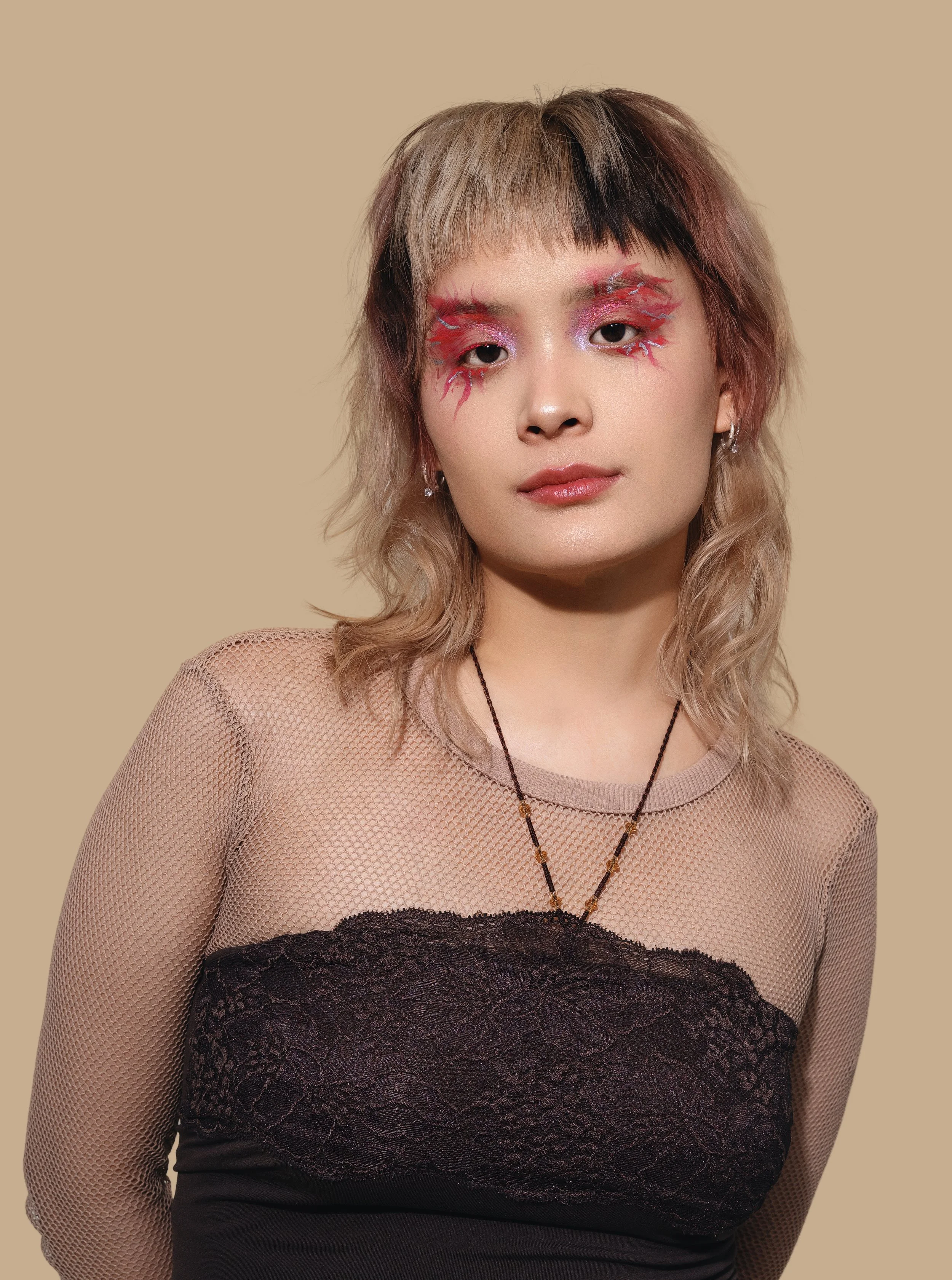 A young woman with pastel pink and black hair and vibrant artistic makeup around her eyes, wearing a beige mesh top with a black lace overlay, and a beaded necklace against a plain beige background.