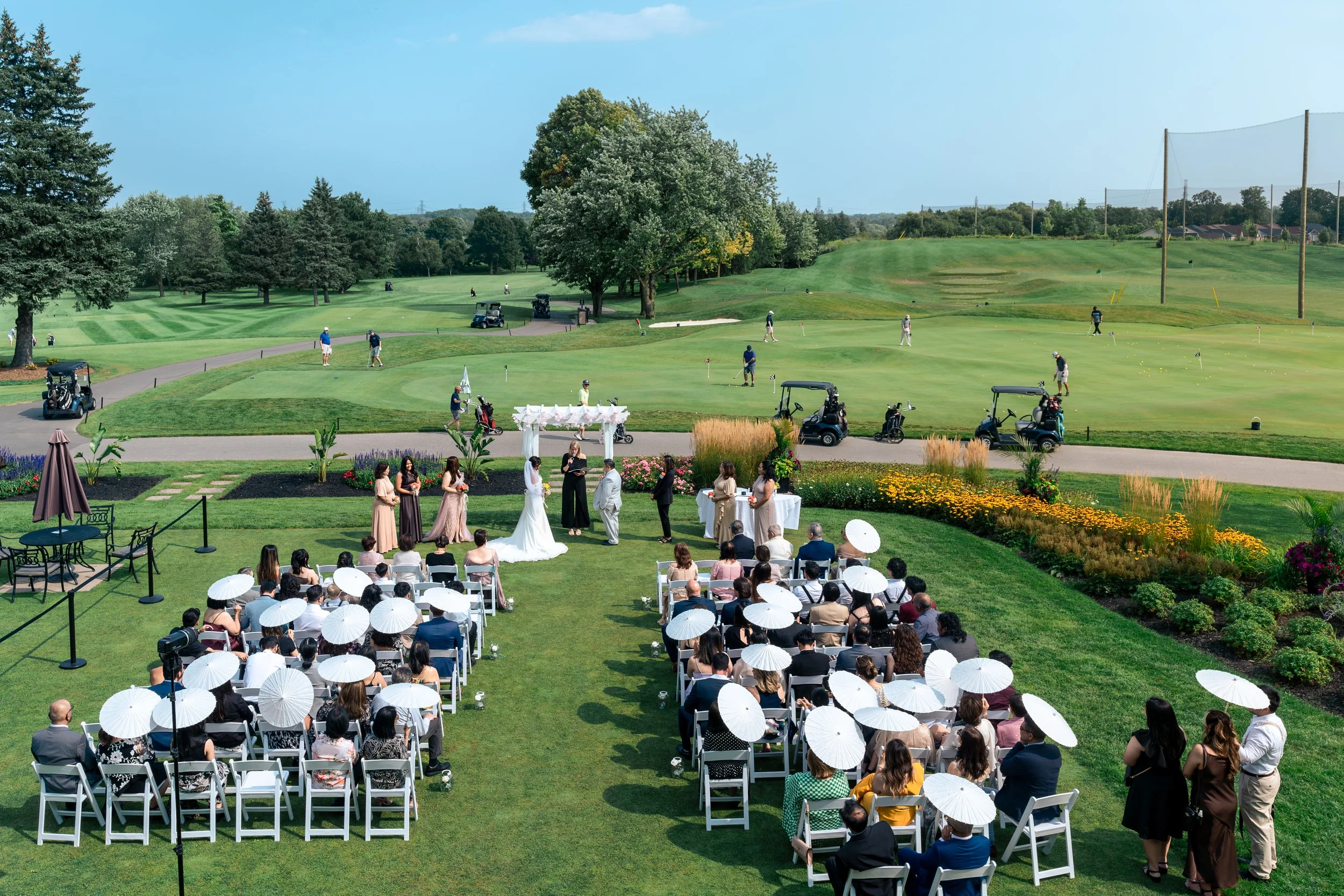 An outdoor wedding ceremony taking place at a golf course, with guests seated under white parasols facing the bride and groom. The ceremony is near lush green grass, colorful flowers, and trees, with players and golf carts in the background.