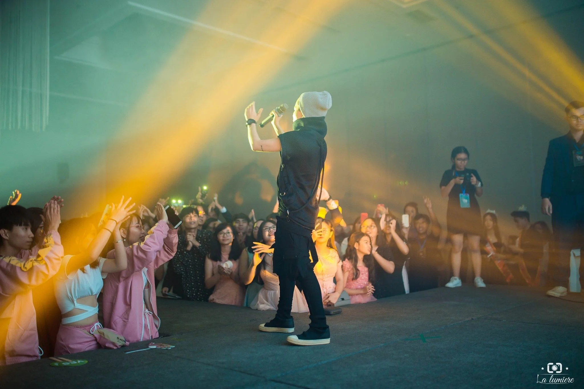 A performer with a microphone on stage entertaining a crowd of fans at a concert. The audience is reaching towards the performer, with some capturing the moment on their phones. Stage lights create a vibrant atmosphere.