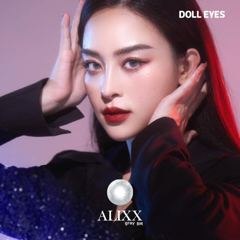 A woman with dark hair and red lipstick poses with her hands framing her face against a pink background, with text indicating "DOLL EYES" and an image of a gray contact lens labeled "ALXX".