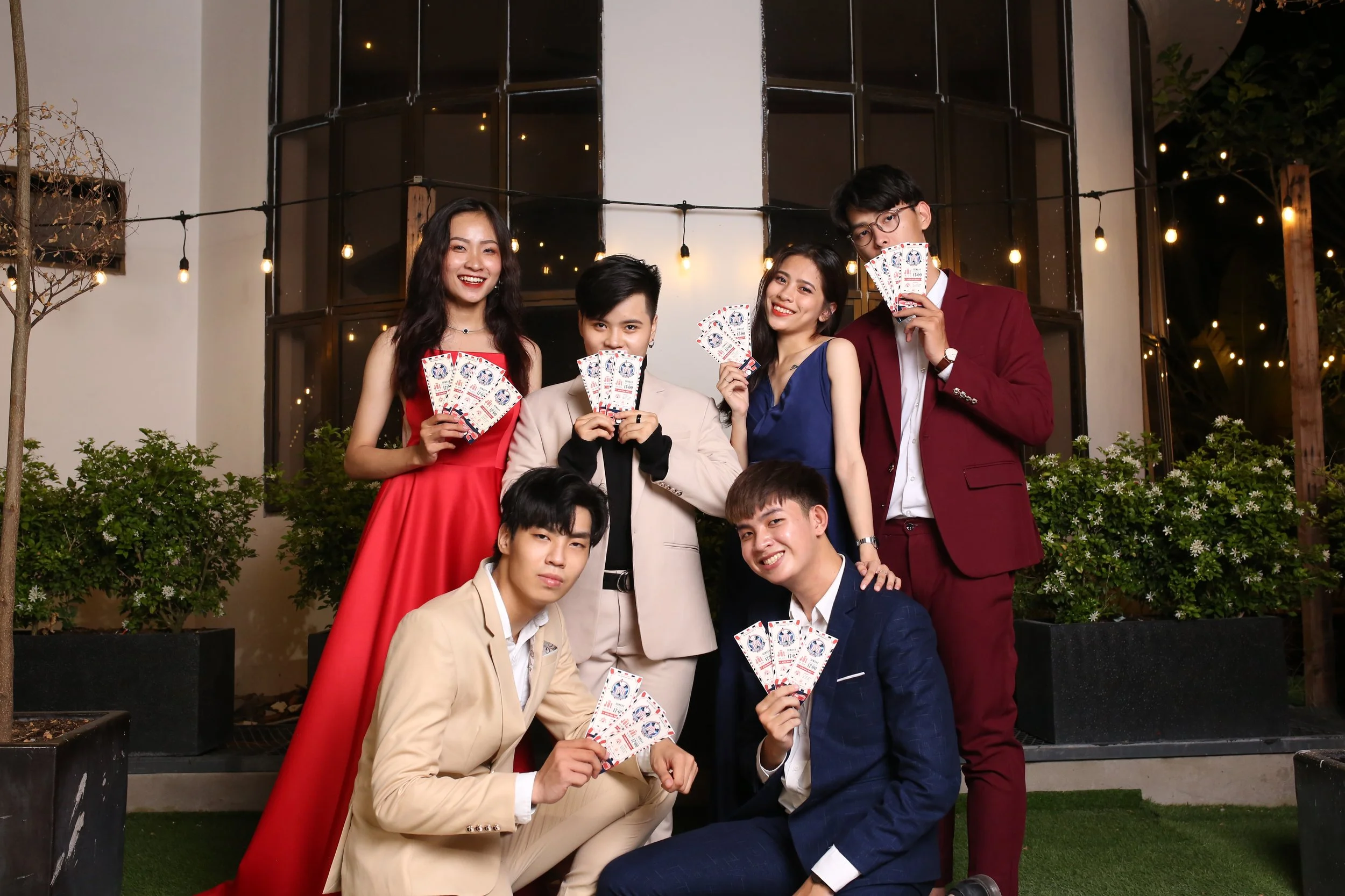 Group of seven young adults dressed in formal attire holding playing cards, standing outdoors in front of large windows and string lights.