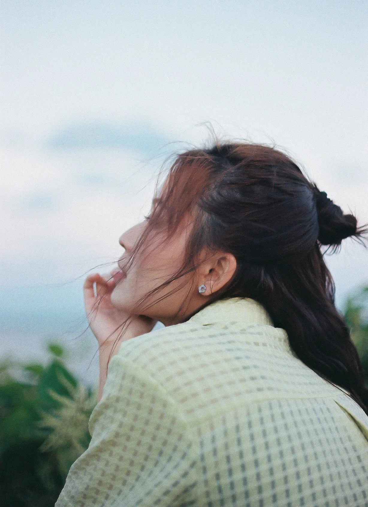 A woman with dark hair and earrings is seen from the side, resting her head on her hand outdoors with a cloudy sky in the background.