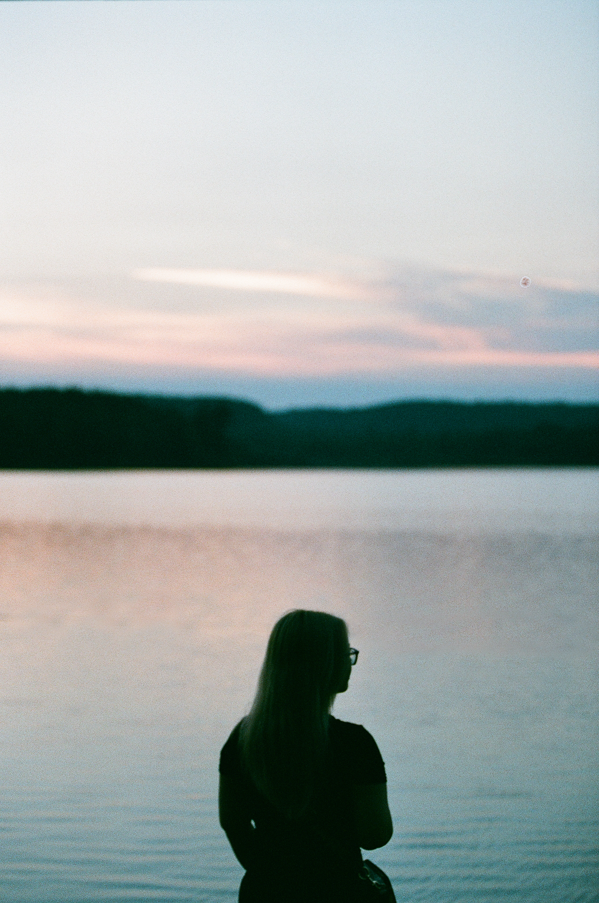 A woman with long hair and glasses standing by a lake during sunset, with mountains in the background.