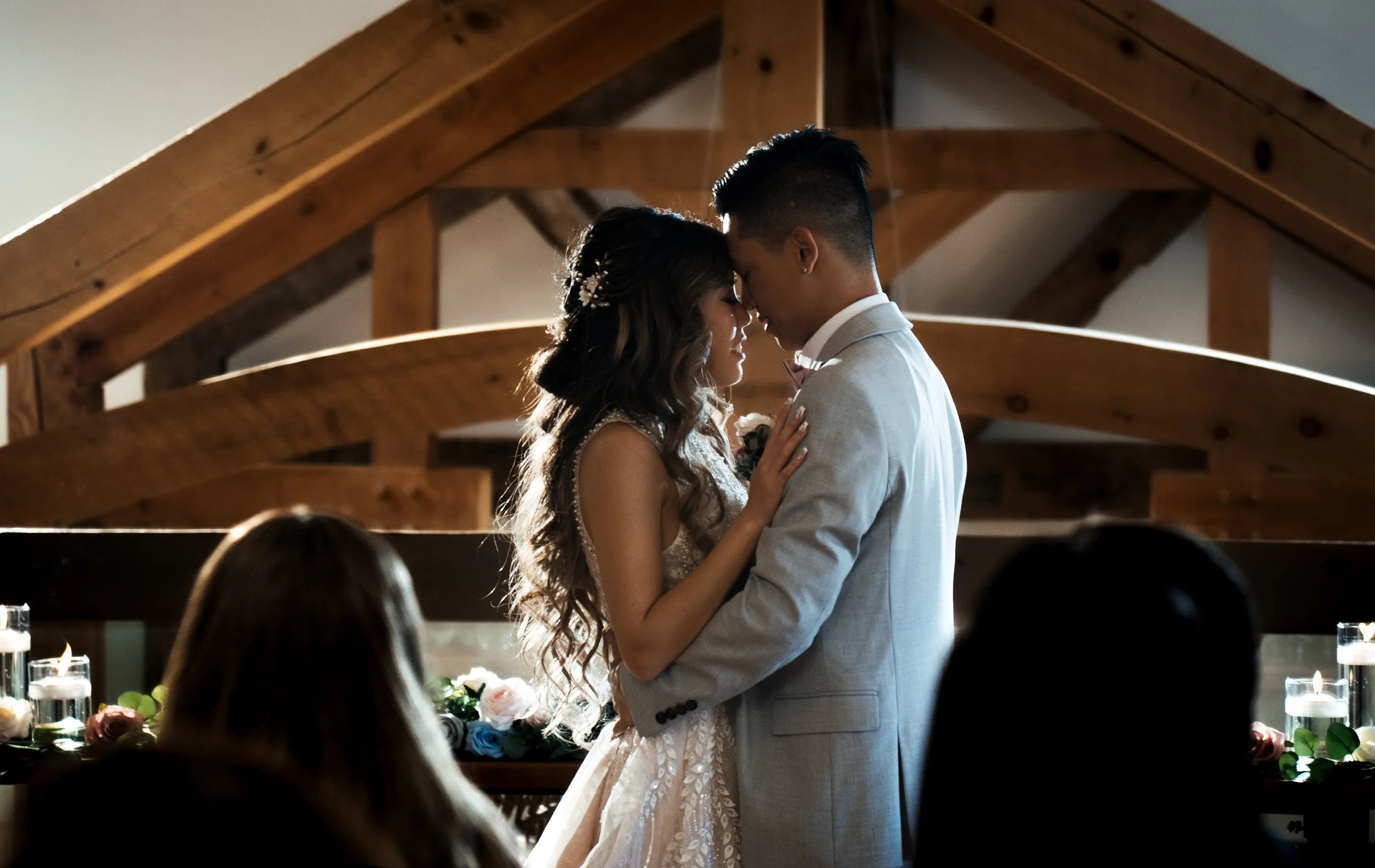 A bride and groom standing close together with foreheads touching during their wedding ceremony inside a wooden venue, with guests watching in the foreground.