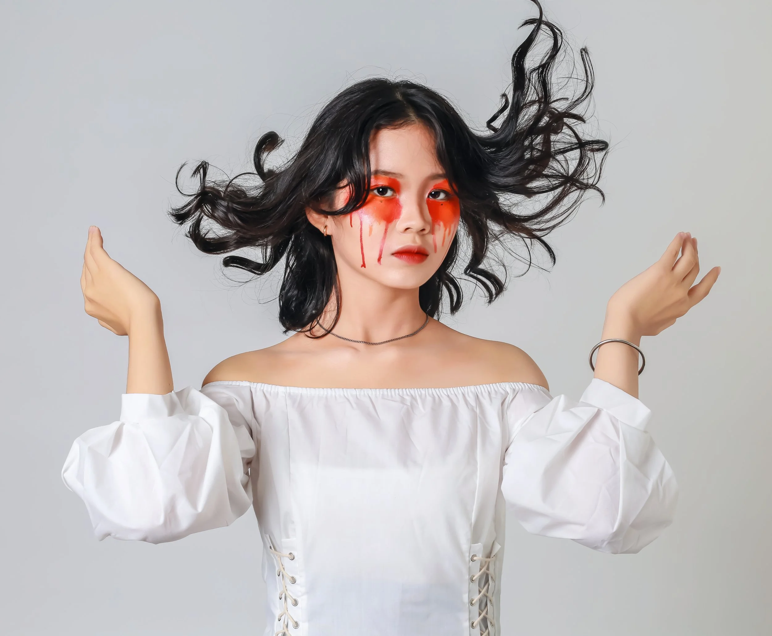 Young woman with black hair and red makeup dripping from her eyes, wearing a white off-shoulder top with lace-up sleeves, posing with her hair flying to the sides against a plain gray background.