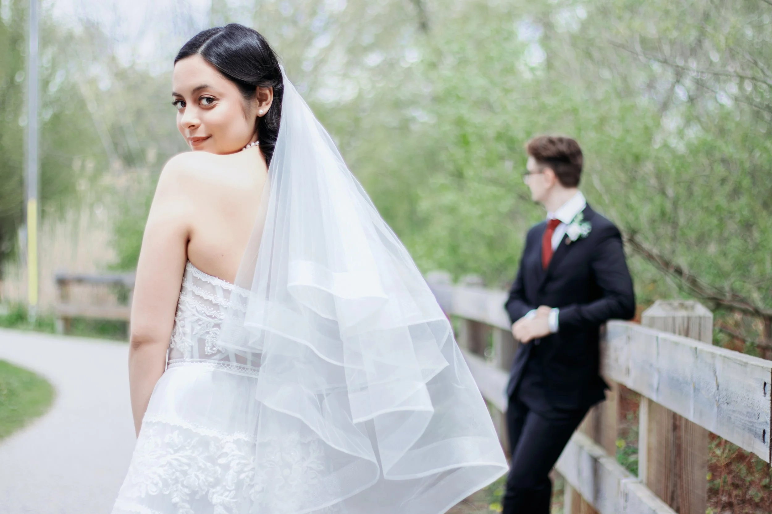 A bride in a white wedding dress with a veil, smiling, standing outdoors on a pathway. In the background, a groom in a black suit with a white shirt, red tie, and glasses, is leaning against a wooden fence and looking away.