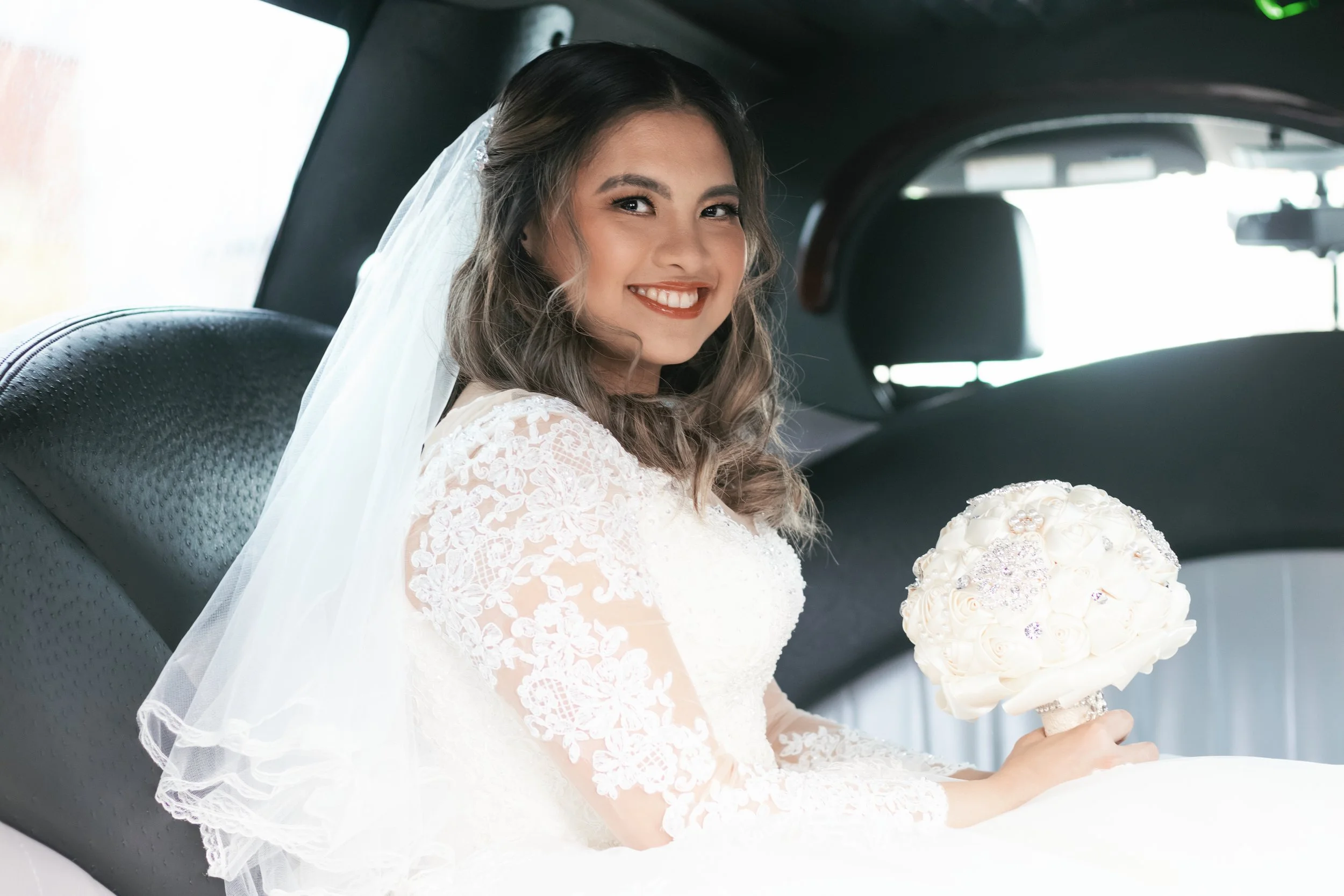 A smiling bride sitting in the backseat of a vehicle, wearing a white lace wedding dress and holding a bouquet of white roses.