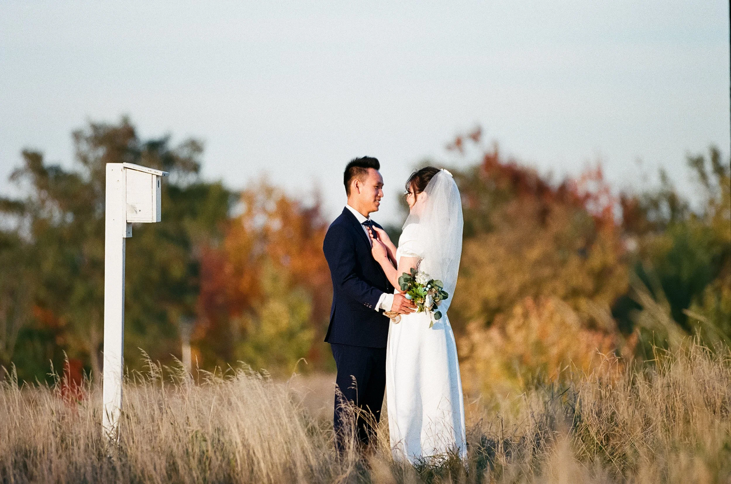A bride and groom standing close together outdoors, smiling and looking at each other, with trees in autumn colors in the background.