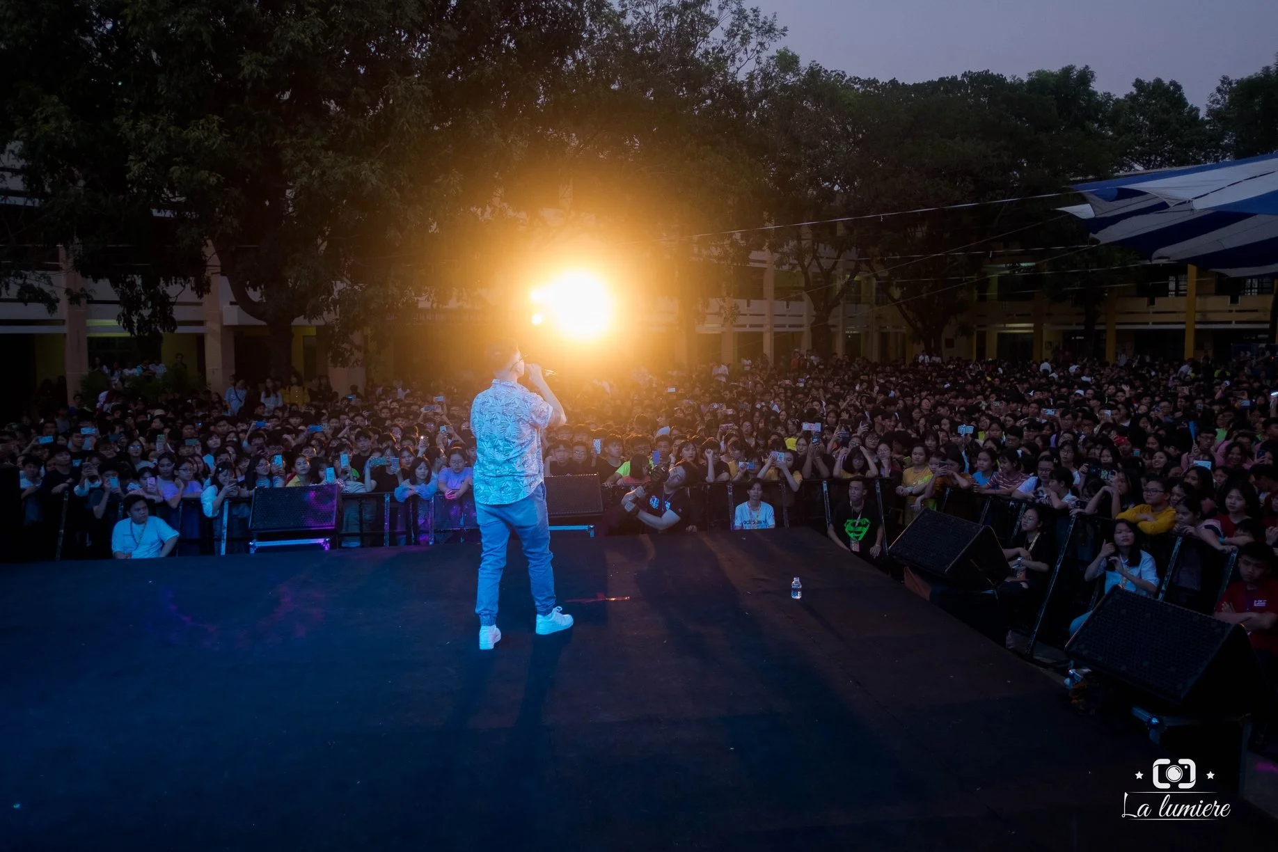A performer on stage with a microphone facing an audience at sunset or early evening, with the sun visible behind the performer. The audience is watching attentively, some holding mobile phones.