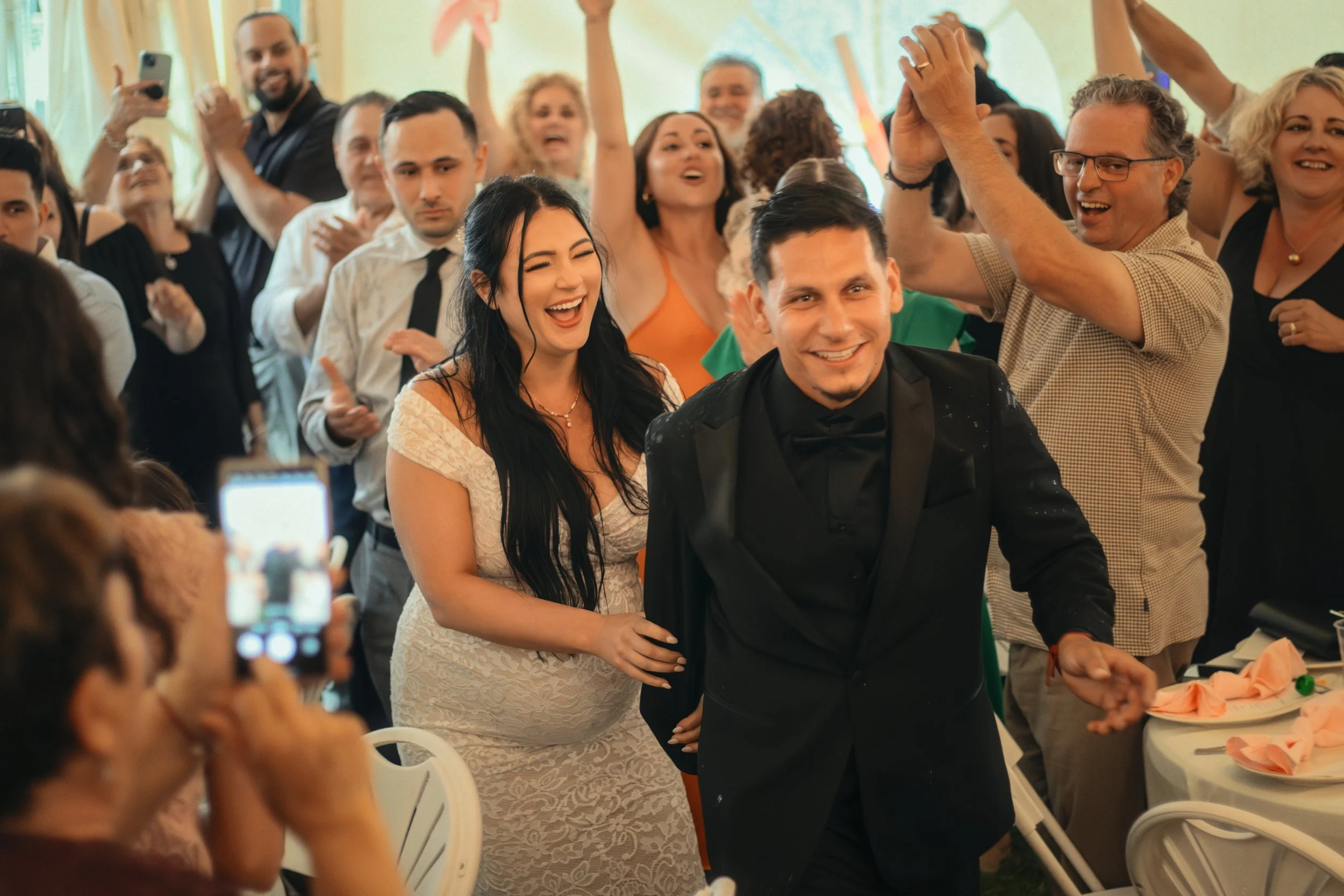 A bride and groom are smiling and walking through a crowd of guests at their wedding reception. The guests are celebrating and taking photos.