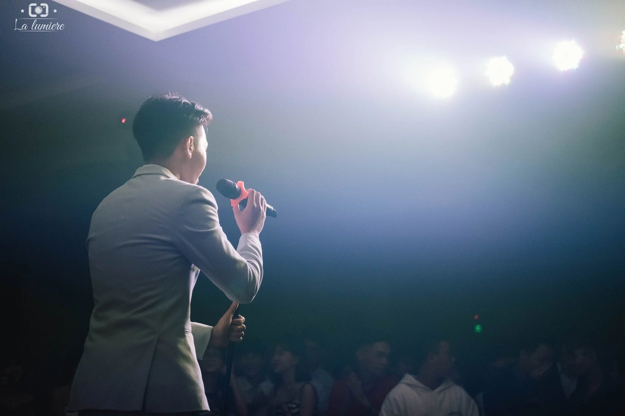 A man in a beige suit holding a microphone, speaking or singing to an audience in a dimly lit room with bright stage lights overhead.
