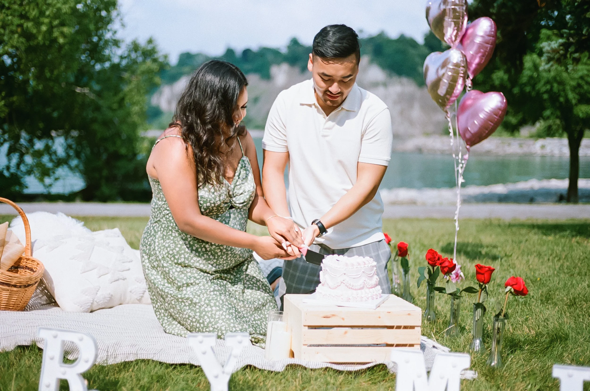 A couple is cutting a pink and white cake at an outdoor celebration near a river, with red roses in vases and pink heart-shaped balloons nearby.