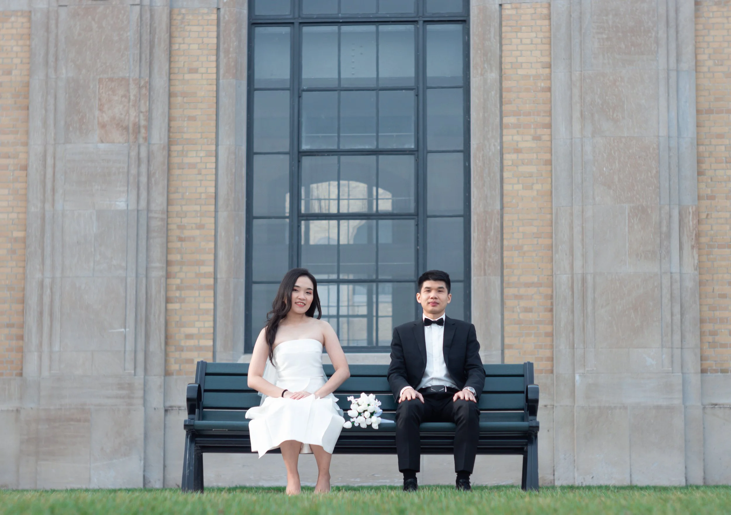 A couple sitting on a park bench in front of a brick and stone building. The woman wears a white strapless dress with a bouquet of white flowers beside her. The man wears a black tuxedo with a bow tie.