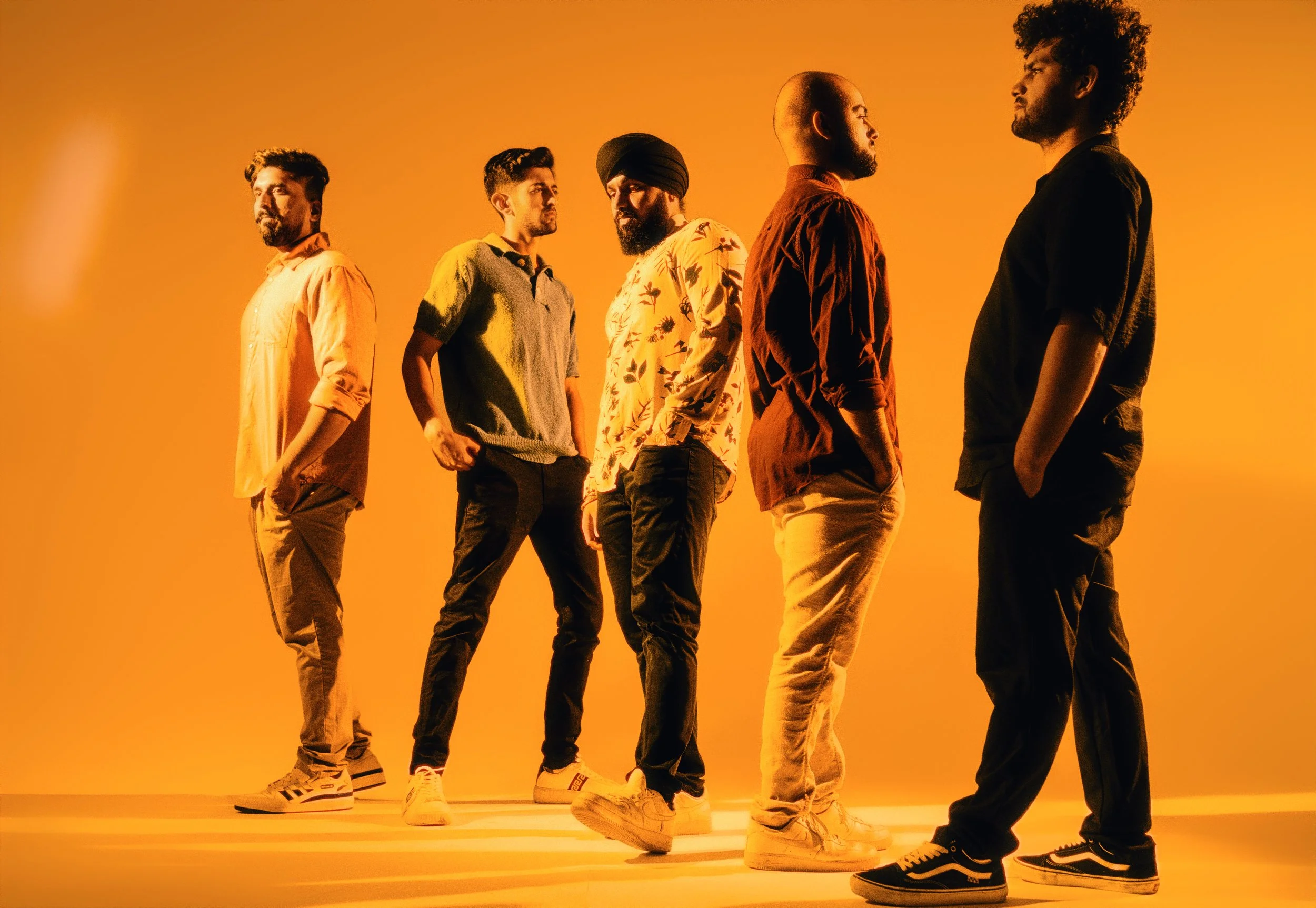 Six men standing in a line against a yellow-orange background, facing each other, dressed in casual clothing, with some with hands in pockets and one with arms crossed, looking serious.