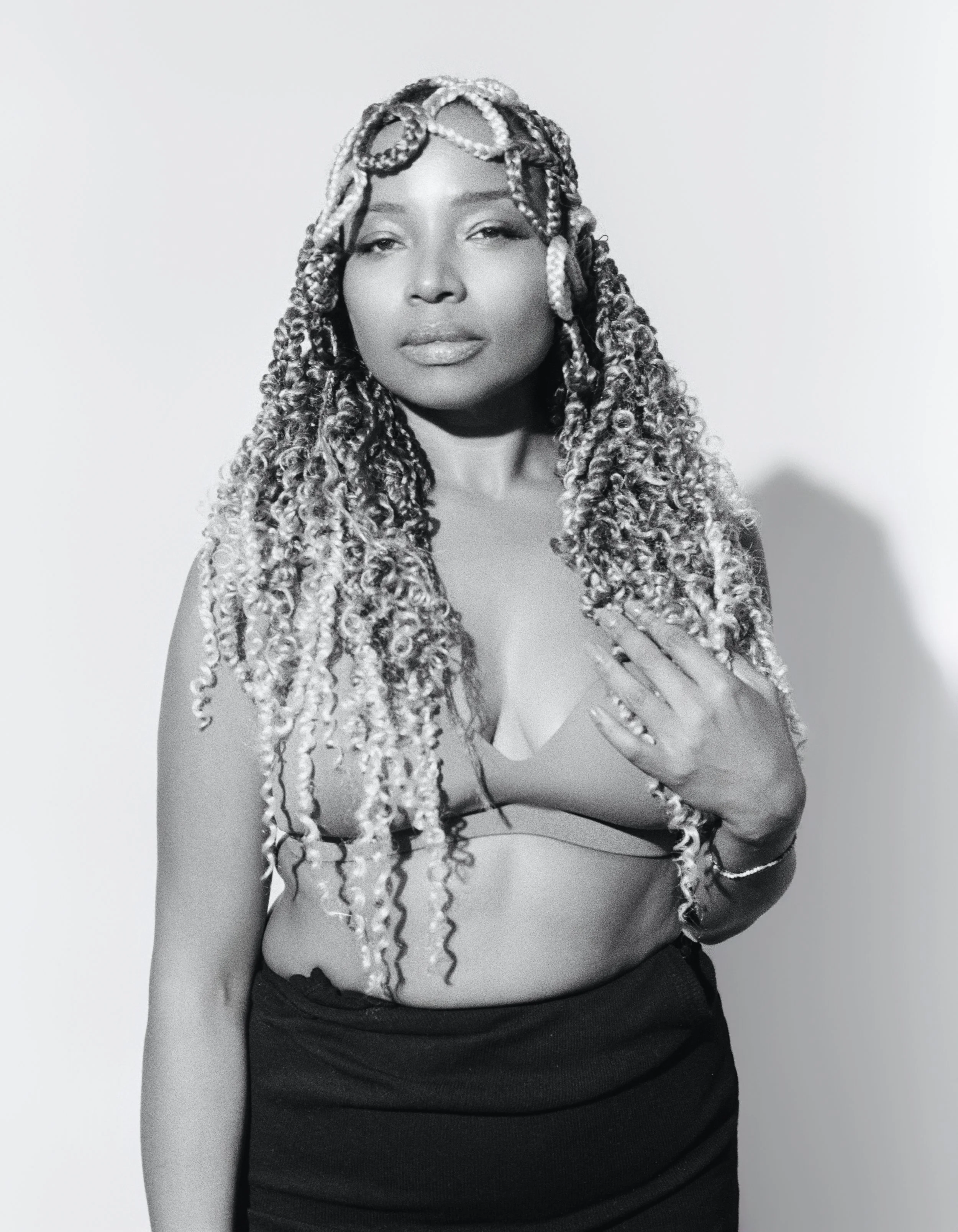 Black and white portrait of a woman with long, curly, braided hair, wearing a sleeveless top and a skirt, standing against a plain background.