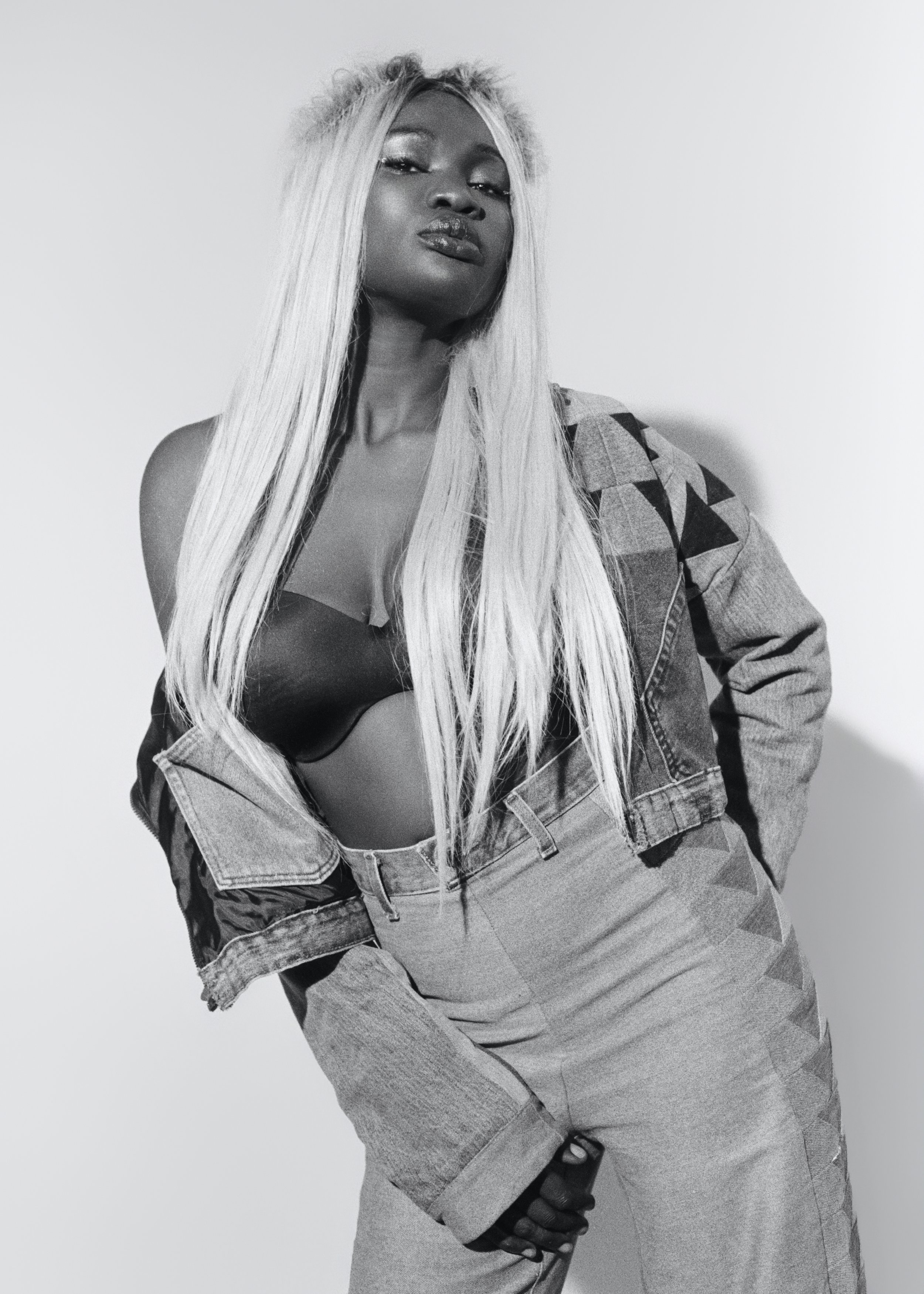 A woman with long, blonde hair poses confidently in a black bra, a denim jacket draped off her shoulders, and high-waisted pants against a plain background.