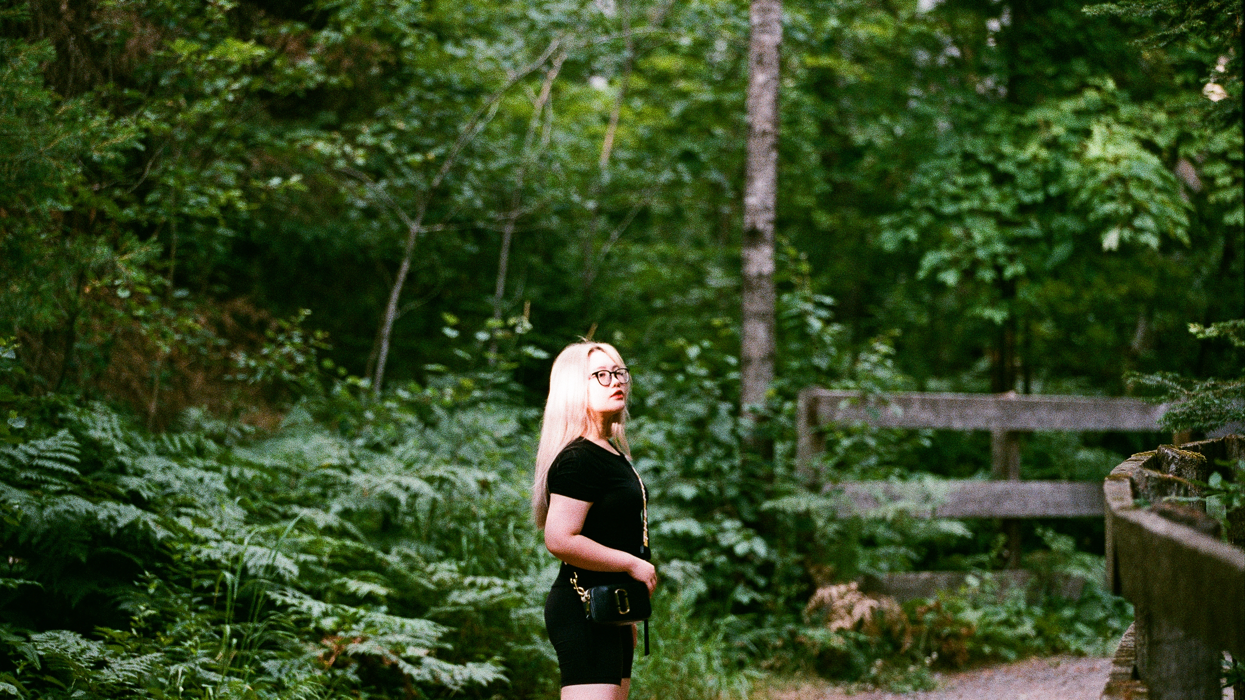 A woman with blonde hair and glasses stands on a forest path next to a wooden railing, surrounded by dense green foliage.