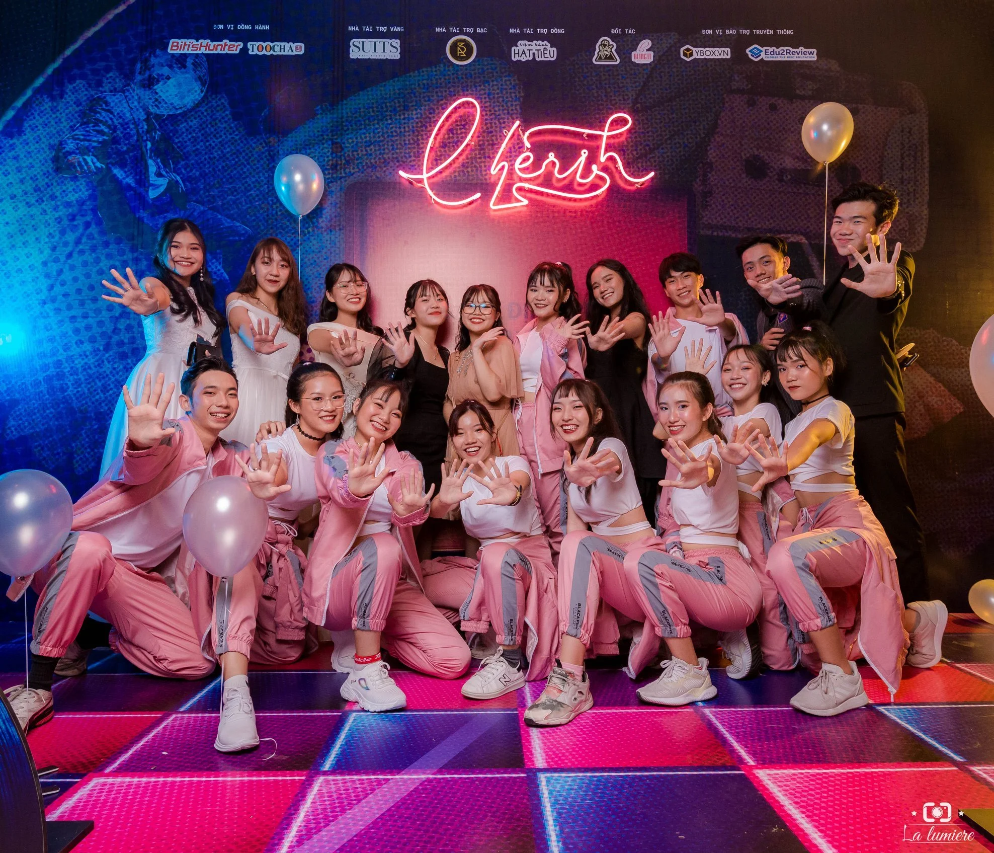Group of young people posing and smiling at a party with neon sign reading 'cherish' in the background, some holding balloons, in a vibrant, colorful setting.