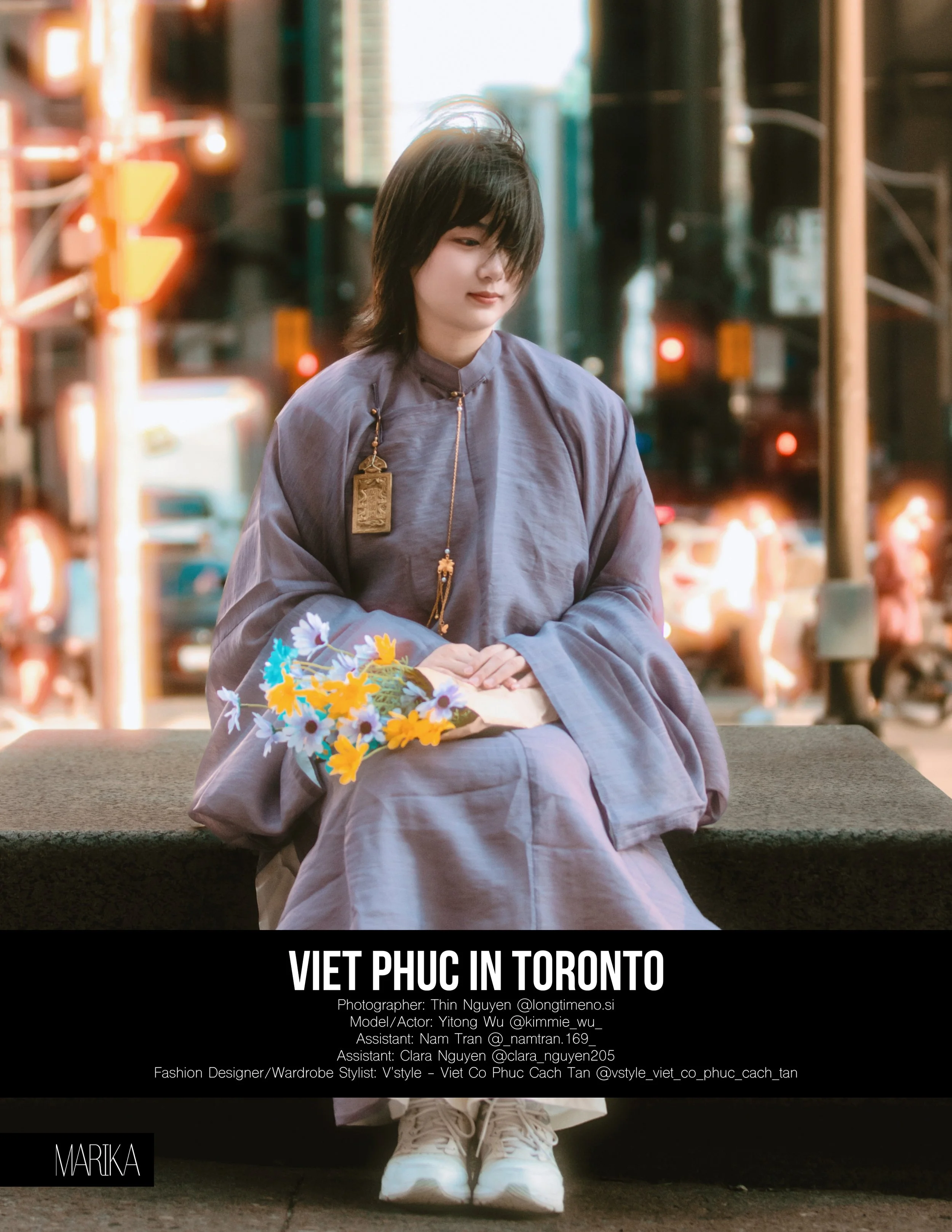 A woman in a gray traditional Vietnamese outfit, sitting on a concrete platform in Toronto, with a bouquet of yellow and purple flowers, and city lights blurred in the background.