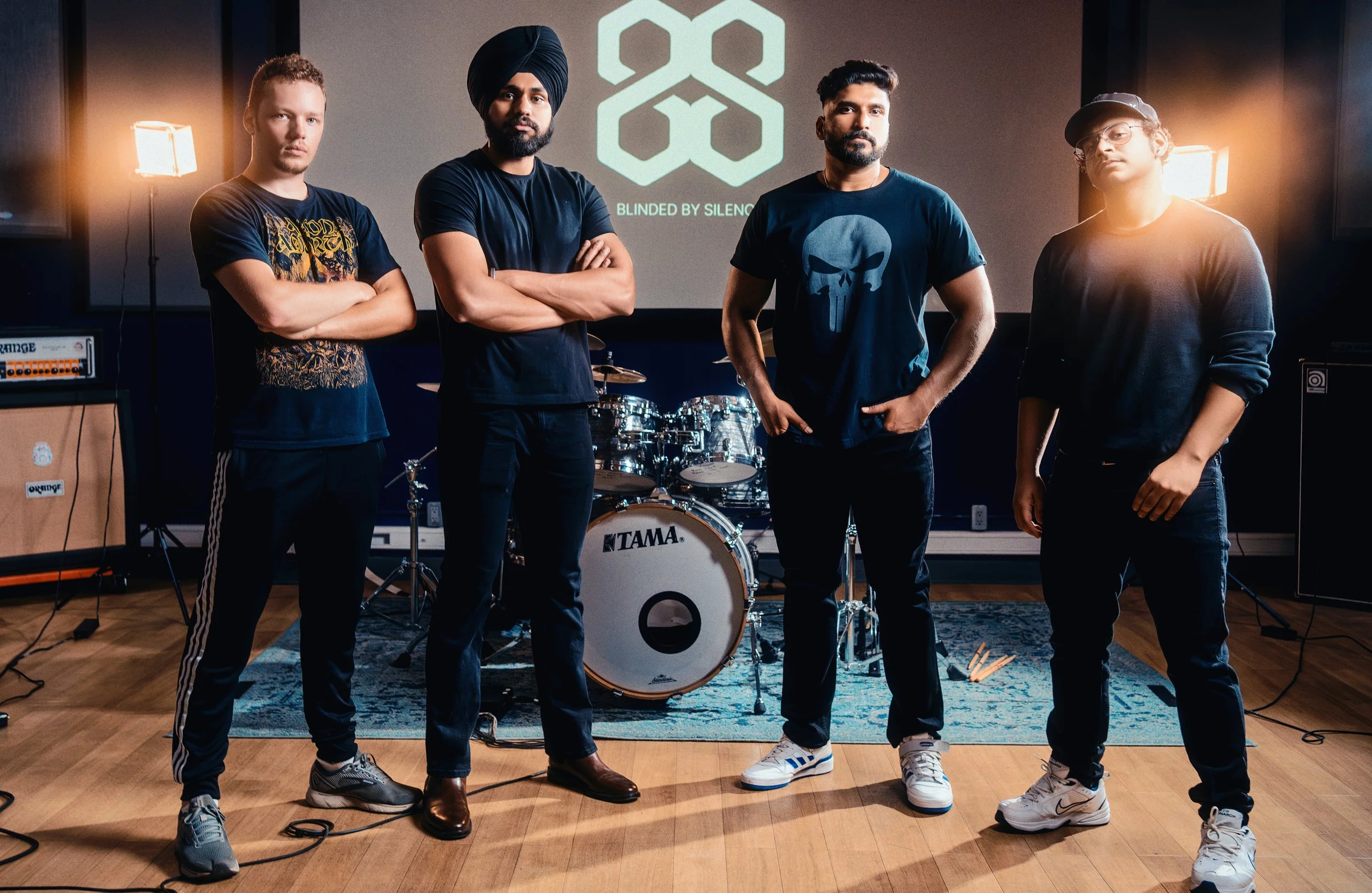 Four men standing in a music studio with musical instruments, stage lights, and a screen behind them displaying a logo and the words "Blinded By Silence."