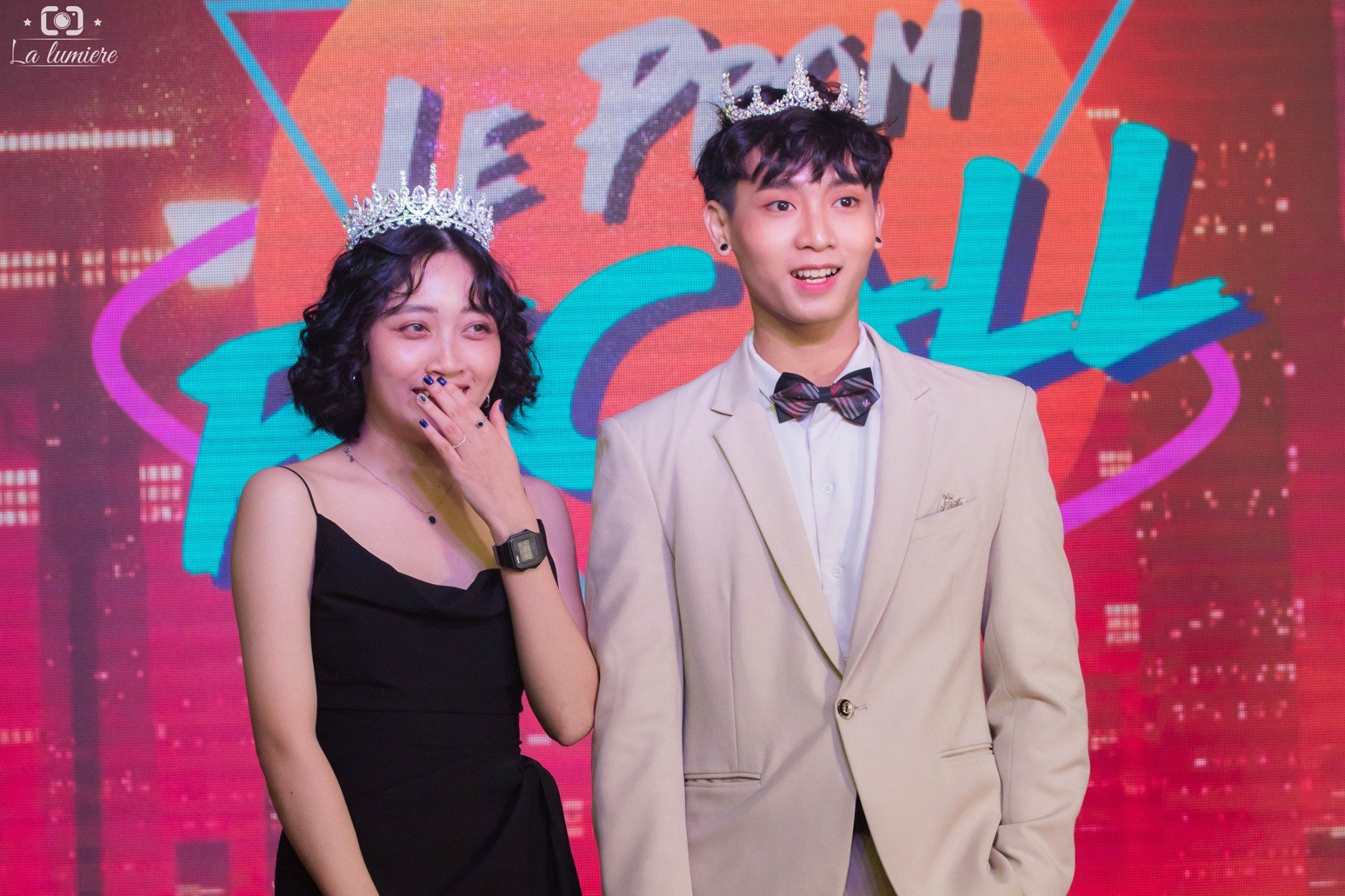 A young woman in a black dress and a young man in a beige suit with a bow tie, both wearing tiaras, standing in front of a colorful background with the text 'LET'S DANCE.' The woman is smiling and covering her mouth with her hand, while the man is sm
