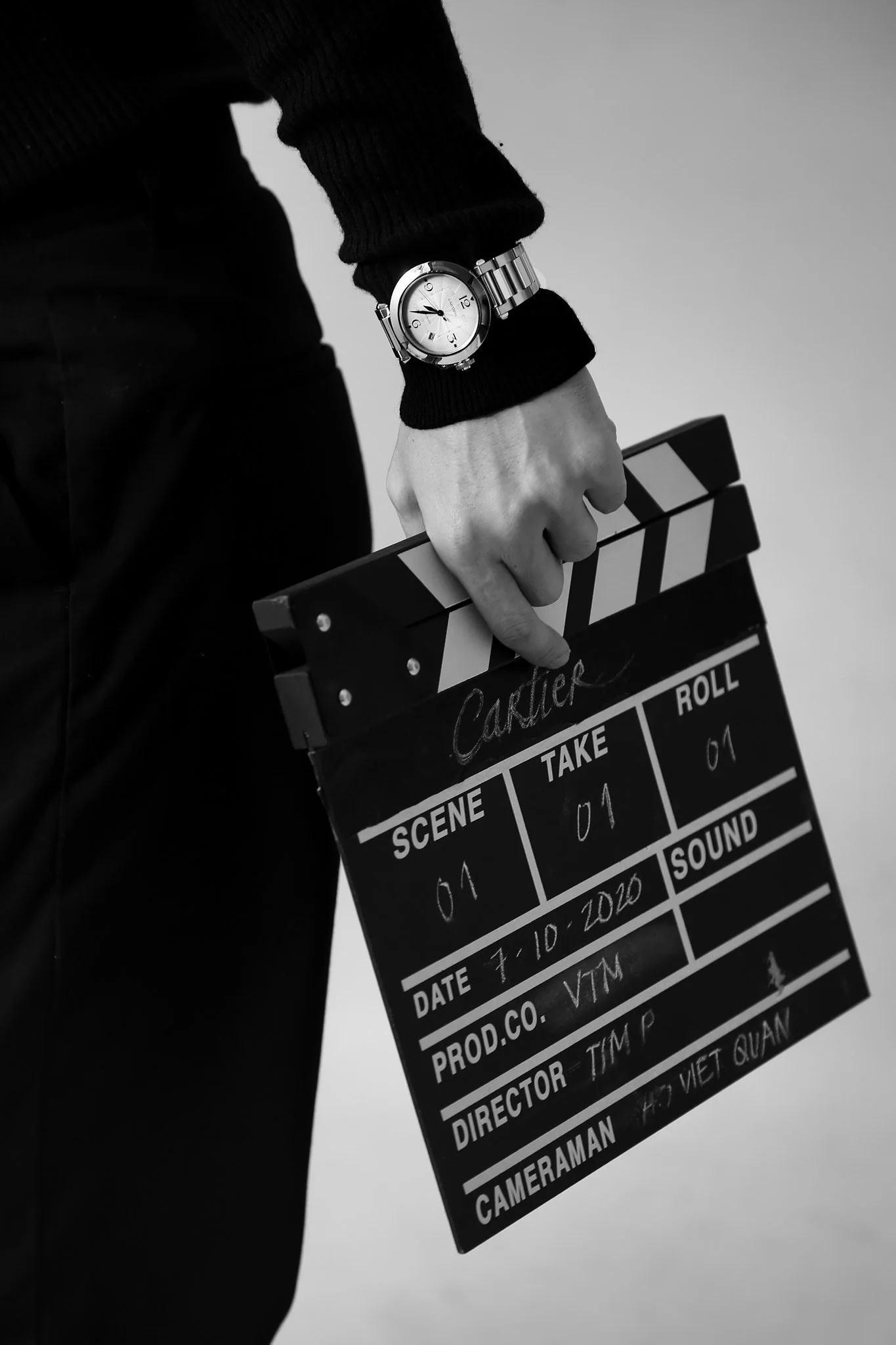 A person holding a black and white film slate with the word 'Cartier' written on it. The slate shows details like scene 04, take 01, roll 01, date 7-10-2020, and other production information. The person is wearing a wristwatch and a black sweater.