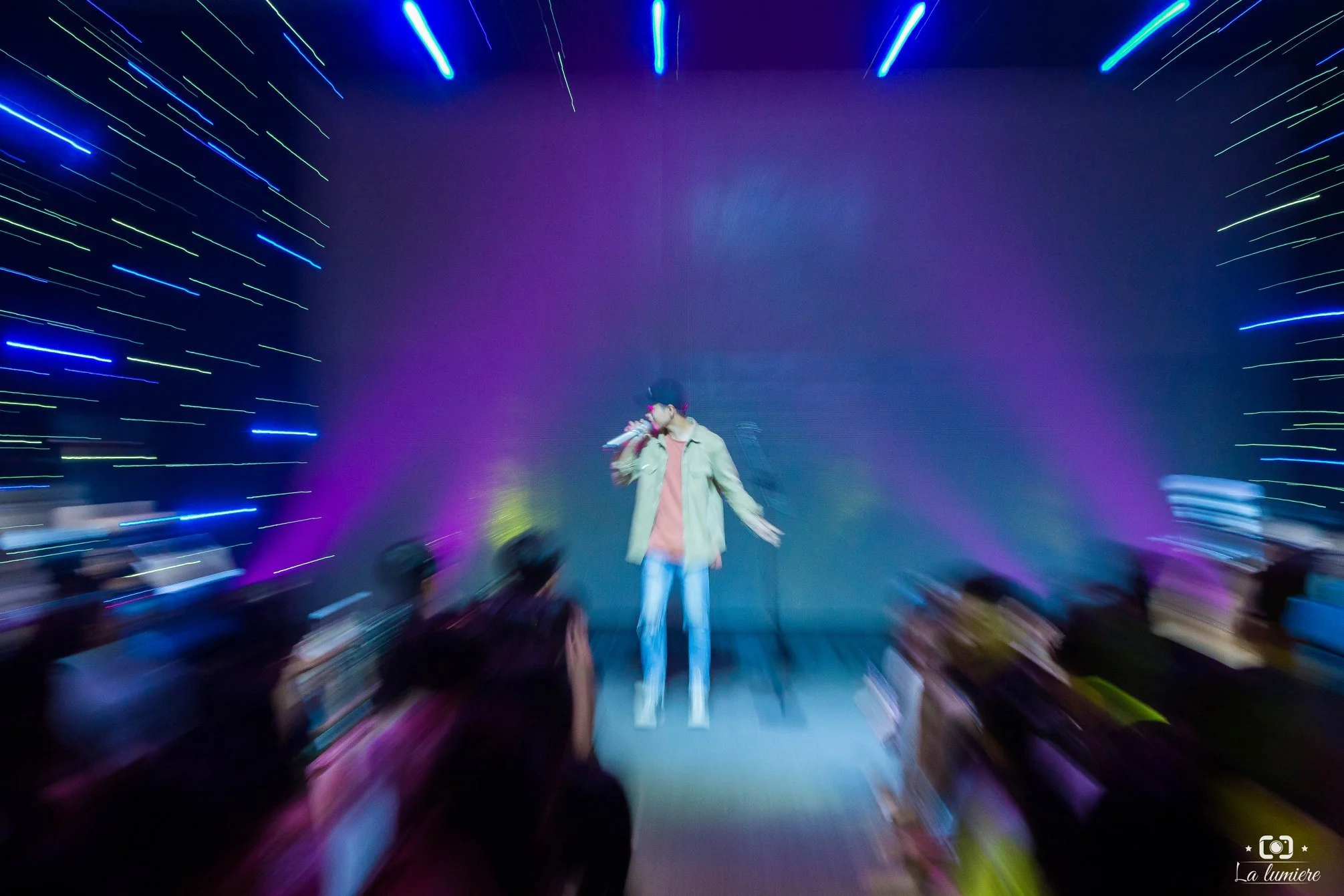 Blurred image of a person singing on stage with colorful lights and an audience in the foreground.