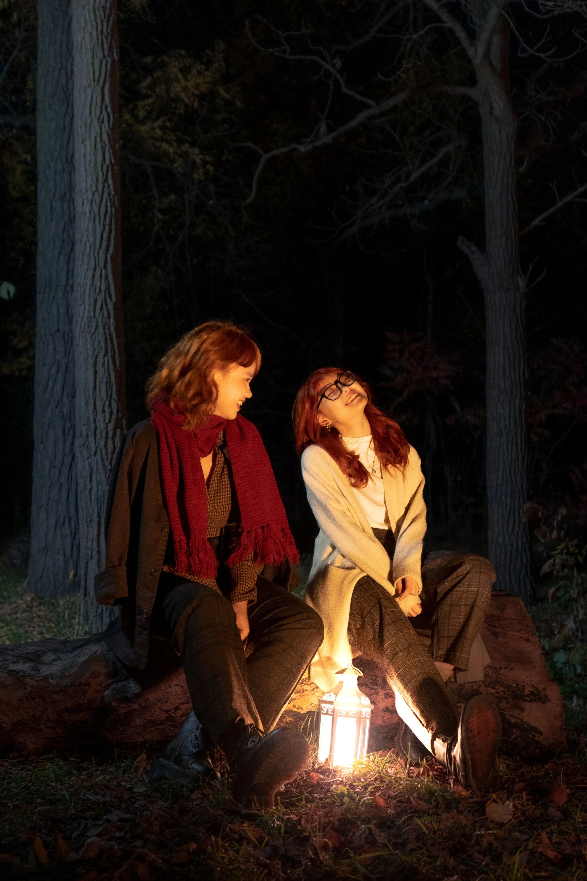 Two women sitting on a large tree trunk outdoors at night, illuminated by a lantern, laughing and enjoying each other's company.