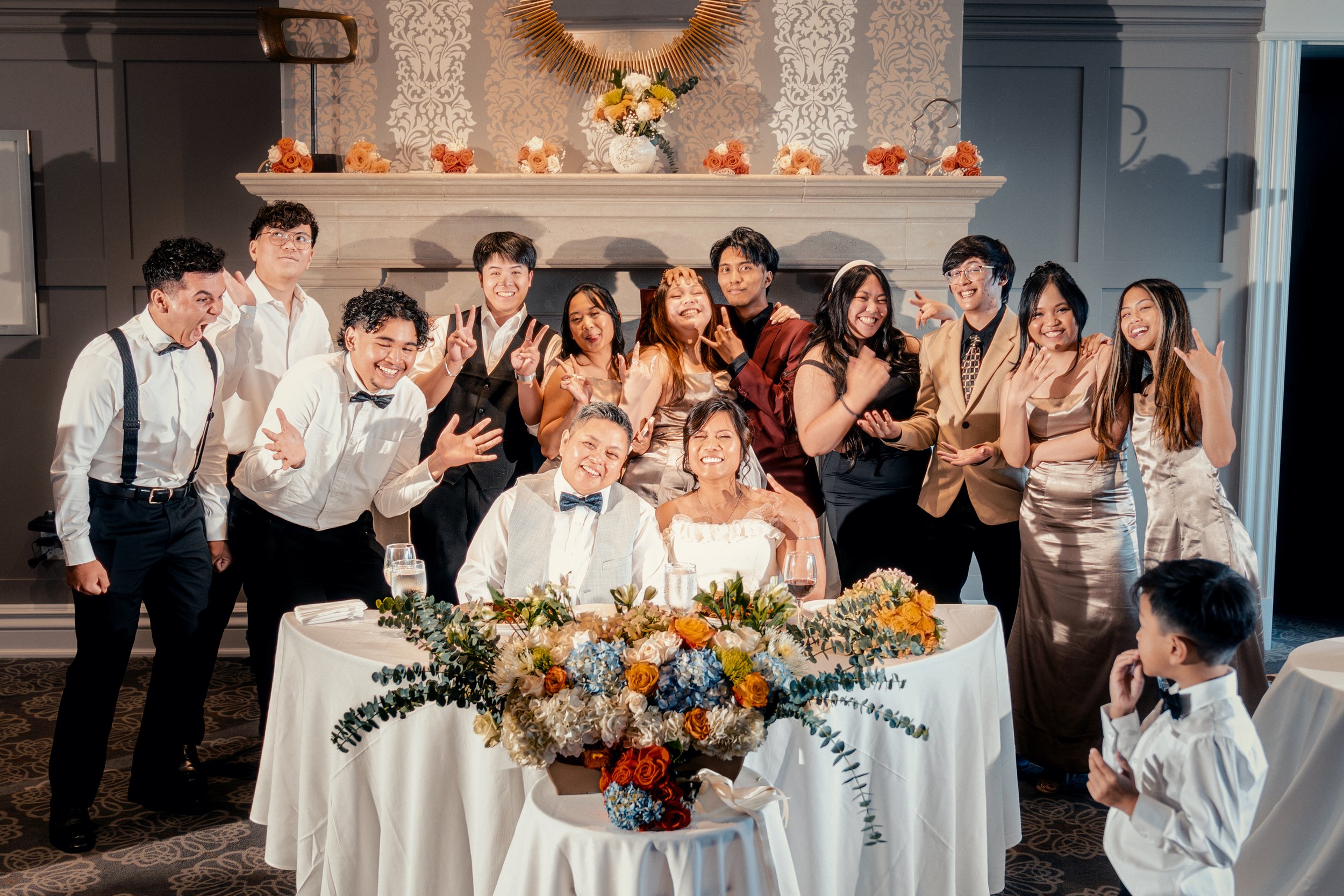 A group of people at a wedding celebration posing for a photo around a table with a large floral centerpiece. The bride and groom are seated at the center, smiling. The group is standing behind them, making gestures and smiling. A young boy is in the