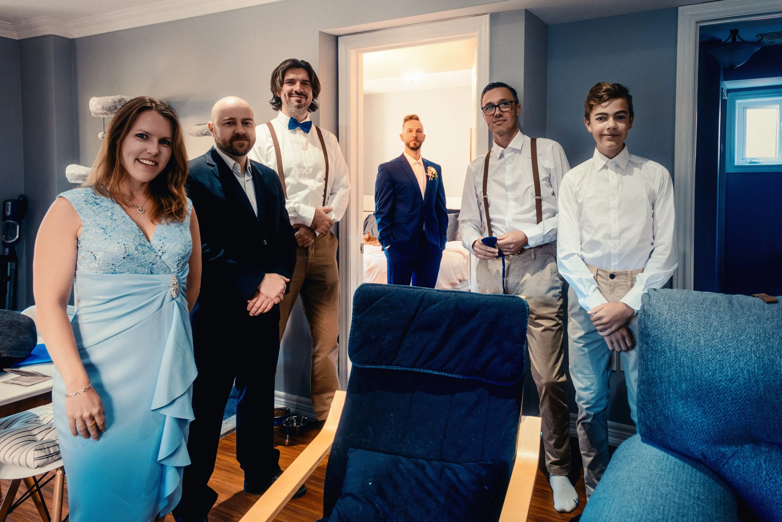 Group of seven people, men and women, dressed in formal and semi-formal attire, standing inside a room with a bed visible in the background, and a doorway leading to another room.