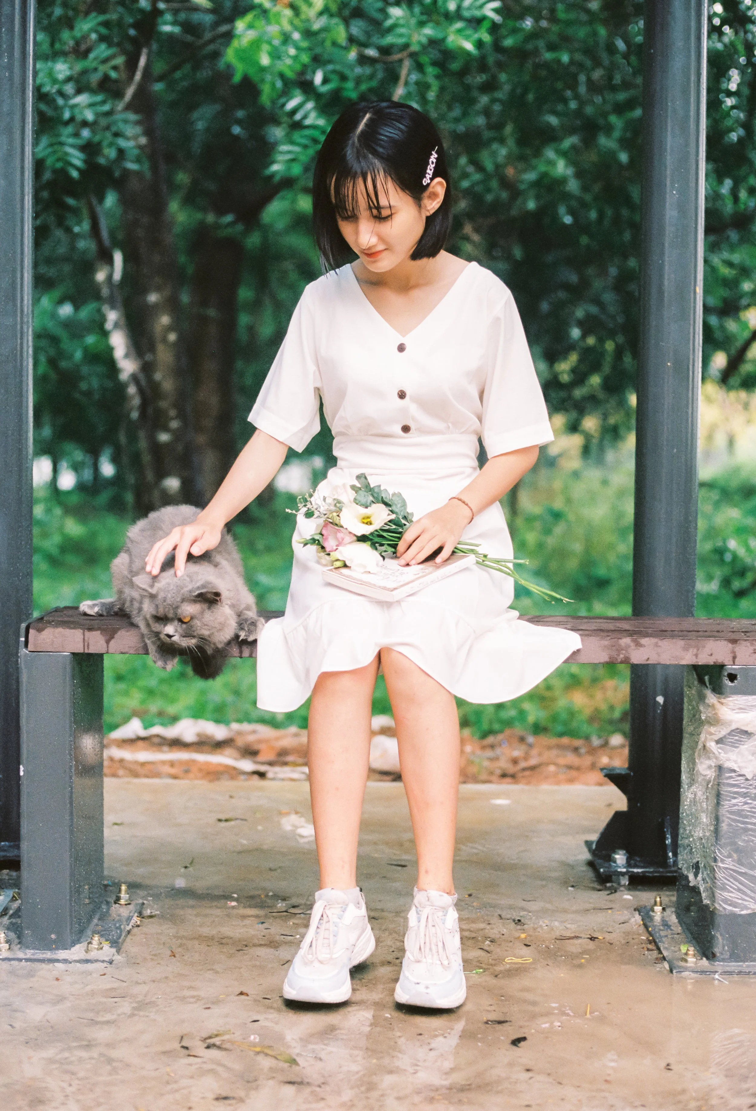 A young woman in a white dress sitting on a park bench, holding a bouquet of flowers, with a gray cat lying beside her on the bench.