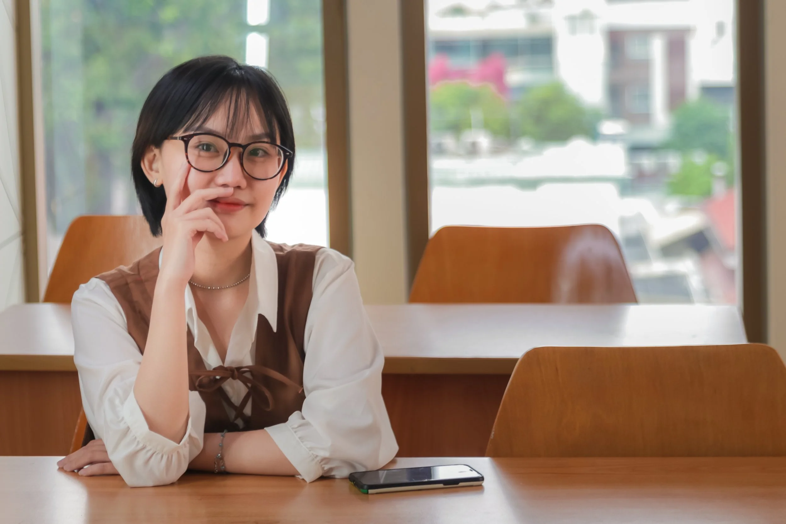 Young woman with glasses sitting at a wooden table in a cafe, looking at the camera, with a smartphone placed in front of her.