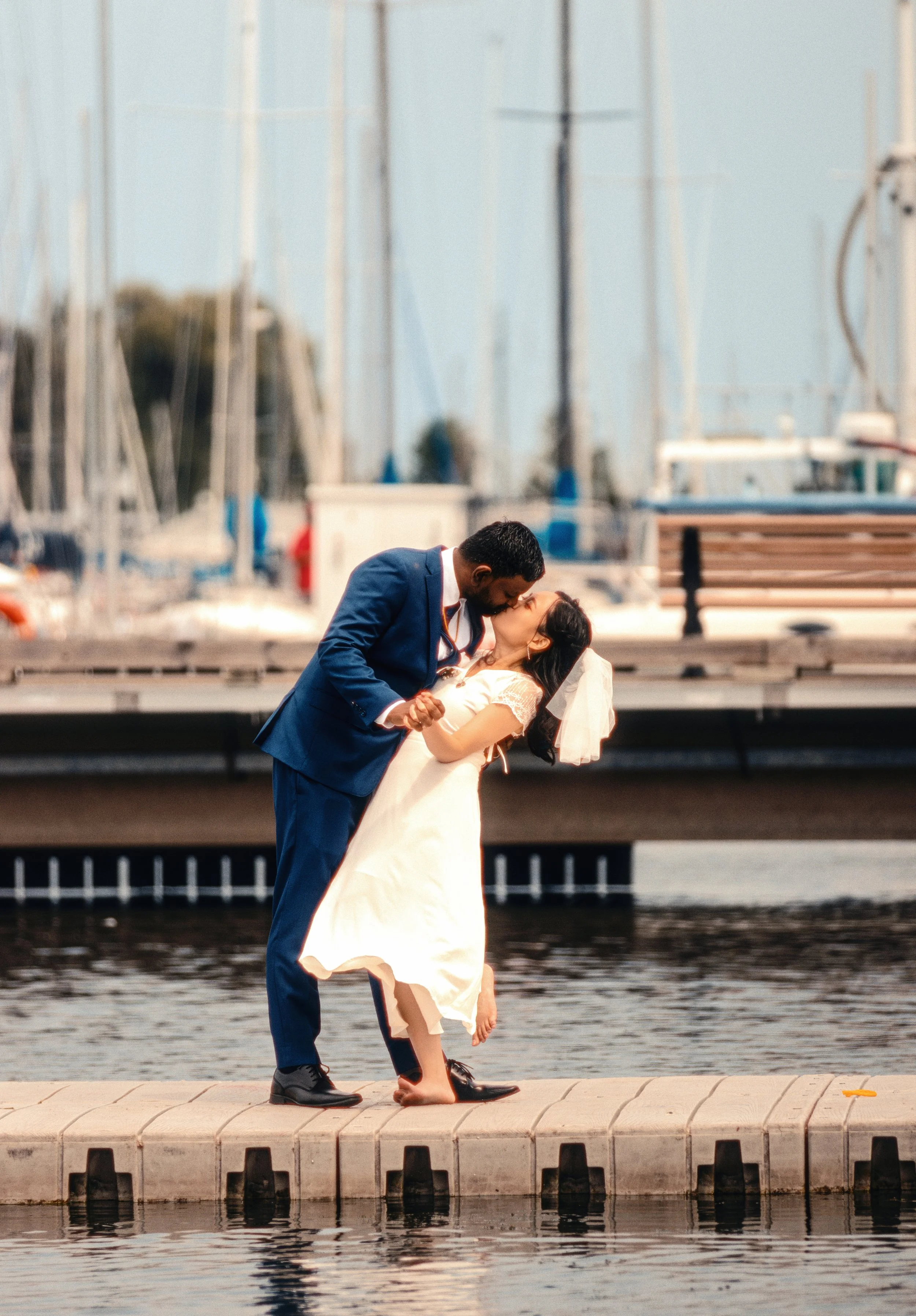 A couple dressed in wedding attire sharing a kiss on a dock by the water, with sailboats and marina in the background.