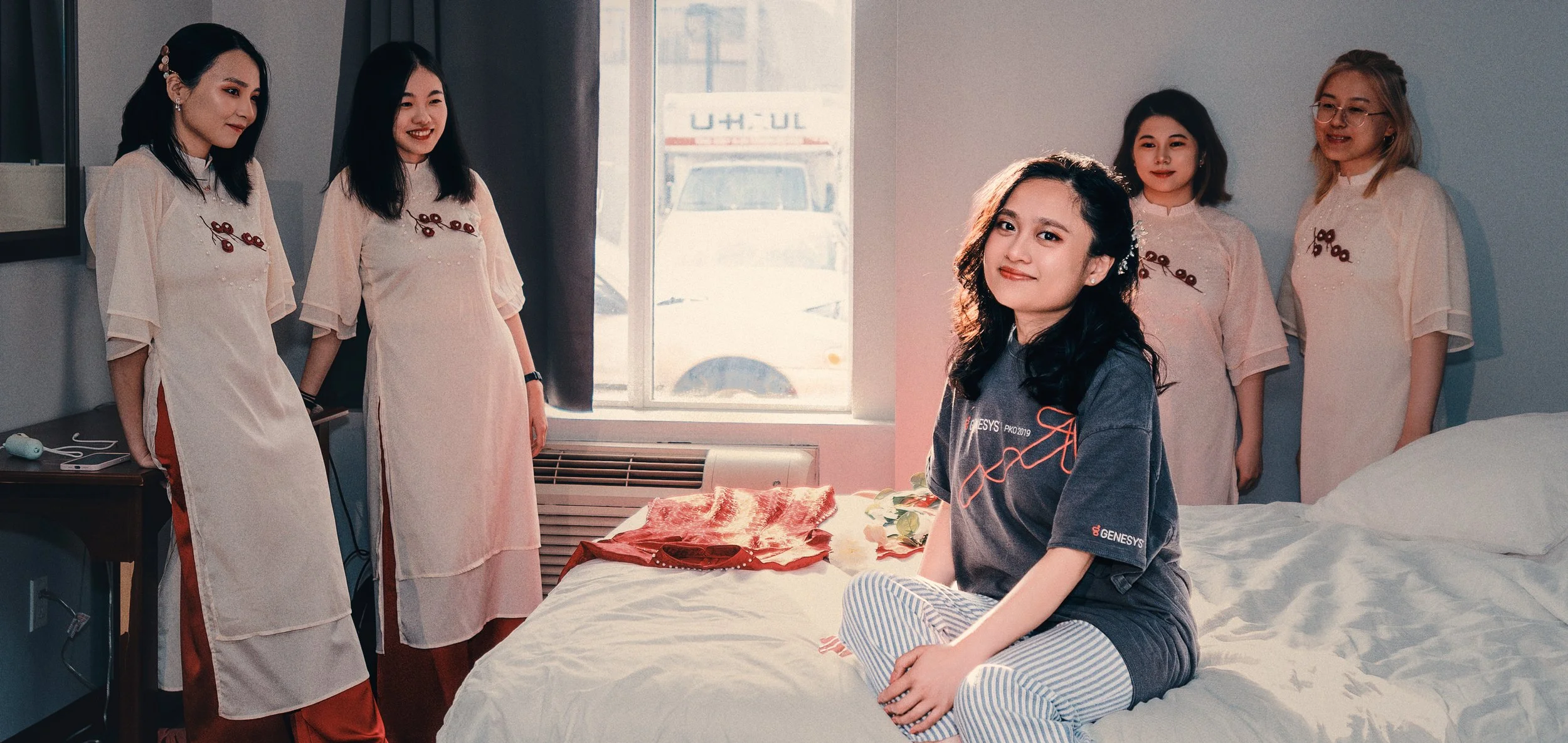 Five women in matching traditional pink dresses with red accents inside a hotel room. One woman, sitting on the bed in casual pajamas, is smiling at the camera. The other four women are standing near the window, chatting and smiling.