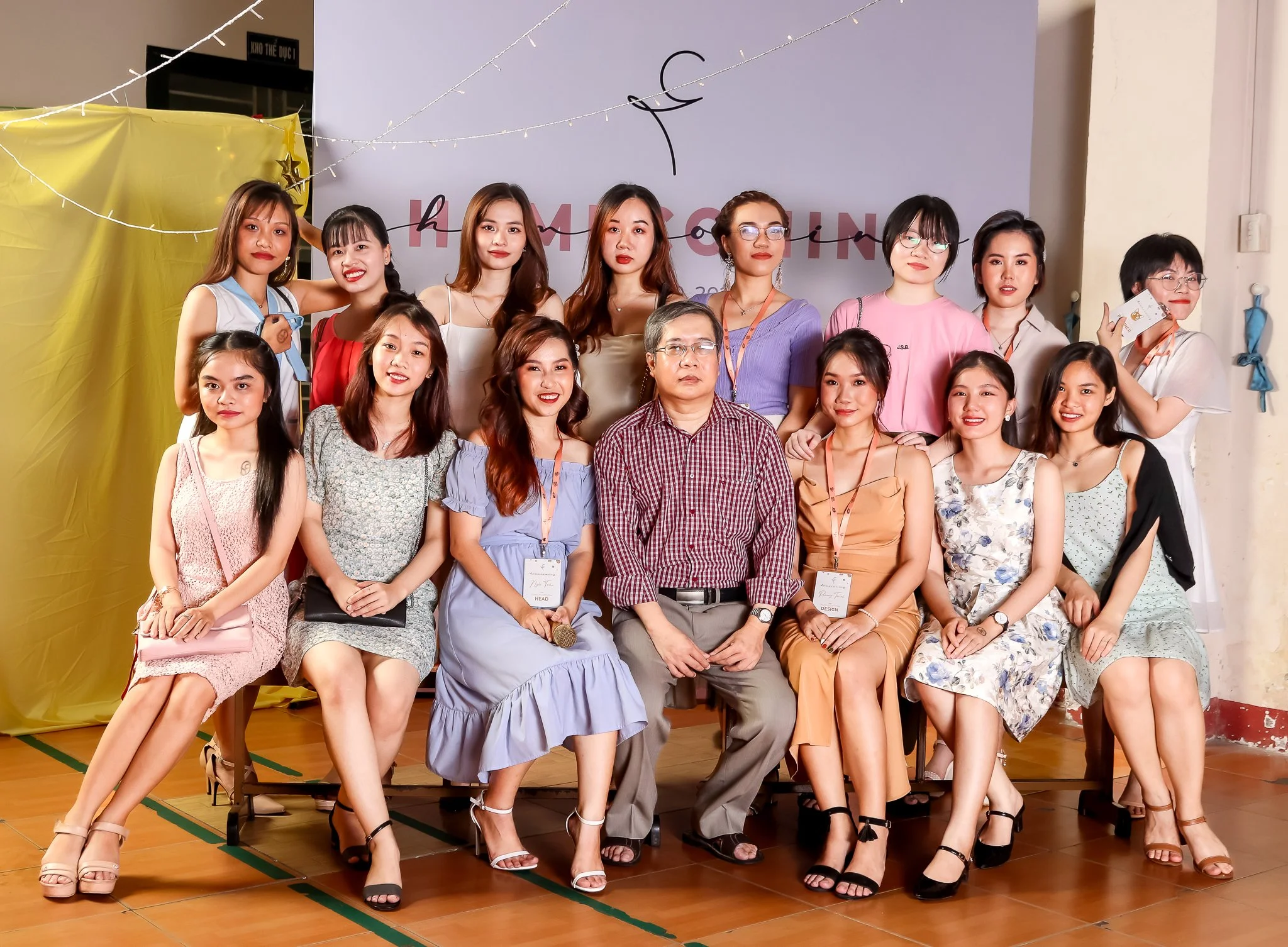 Group of women and a man posing for a photo during an event, with a backdrop that has the words 'HƯM GẮNG' on it, in an indoor setting with string lights and decorations.