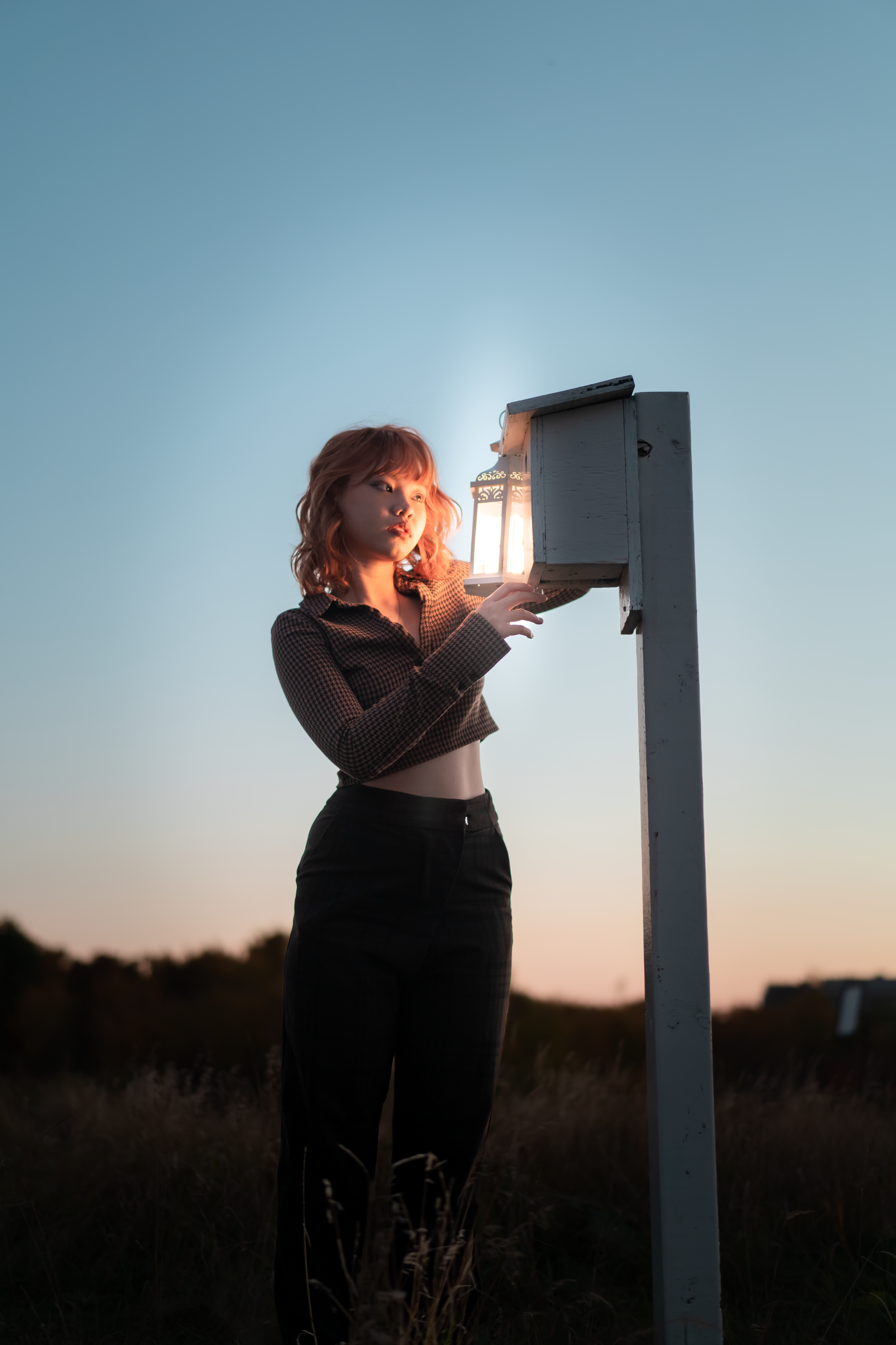 A young woman with red hair and a checkered shirt stands outdoors at dusk, examining a small lantern mounted on a wooden post.