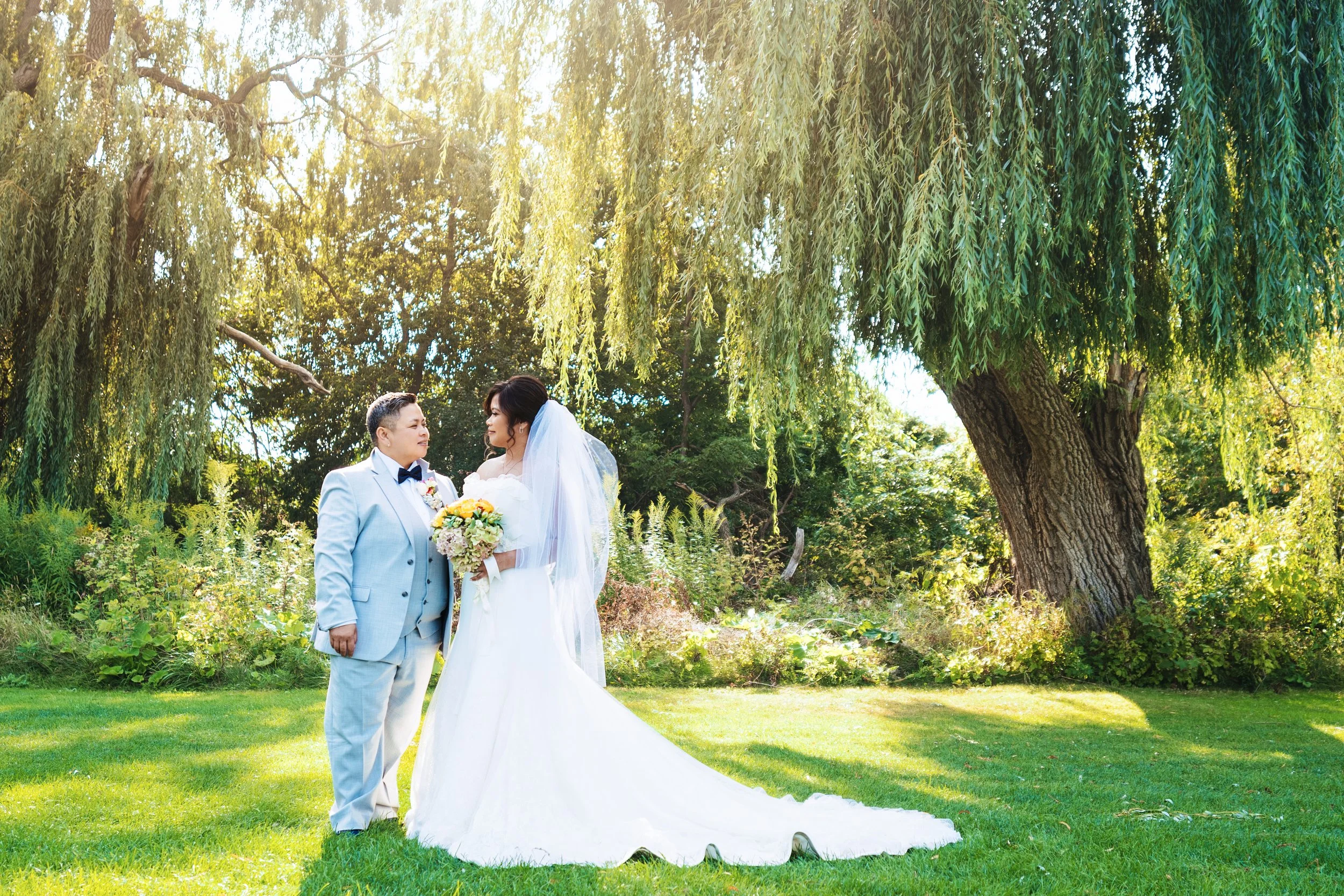 A bride and groom standing together outdoors on a sunny day, under a large tree with hanging branches, surrounded by green grass and foliage, sharing a moment during their wedding.