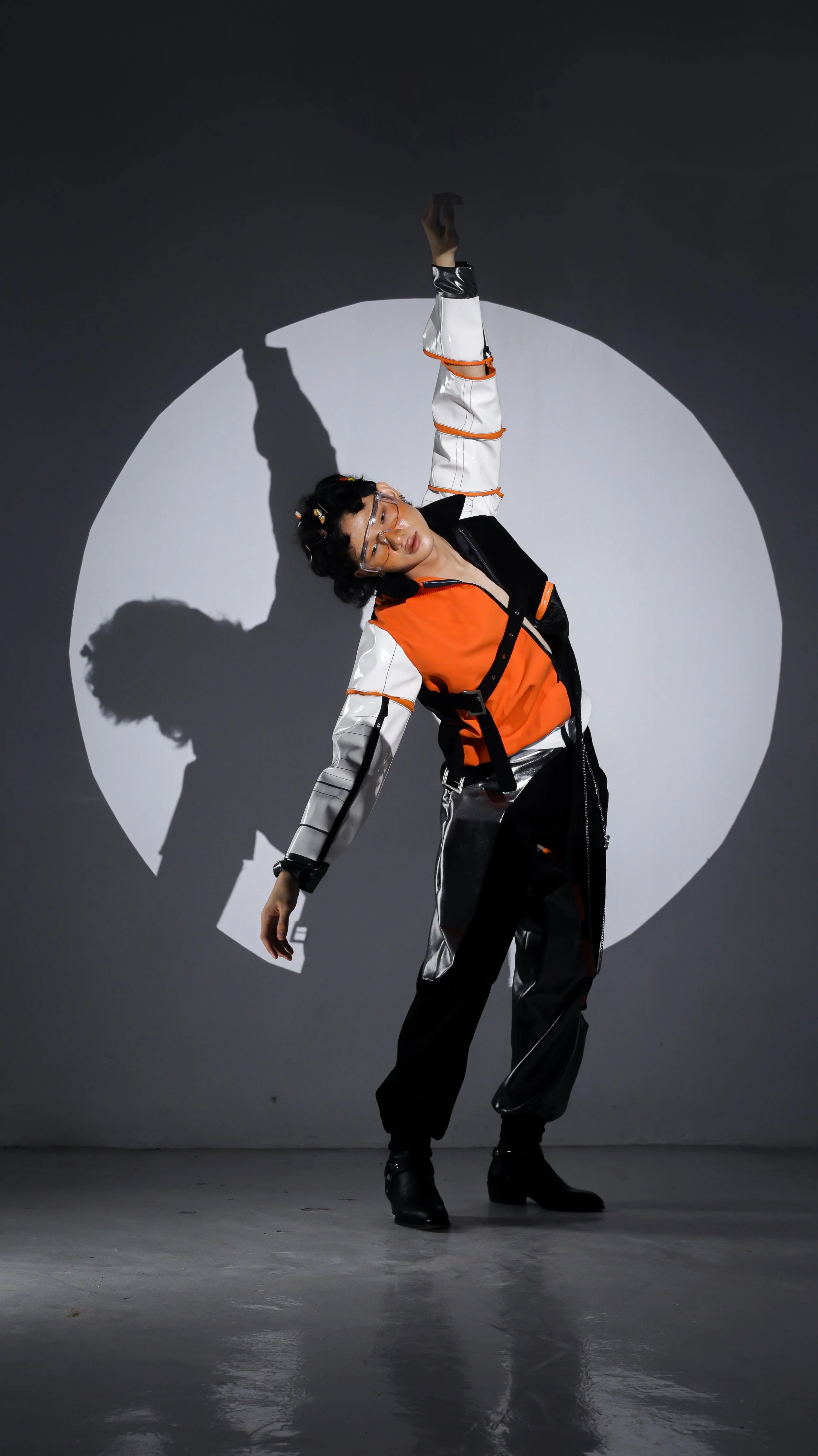 A person posing with one arm raised over their head, dressed in a futuristic orange, black, and silver outfit, standing against a gray background with a white circle and shadow.