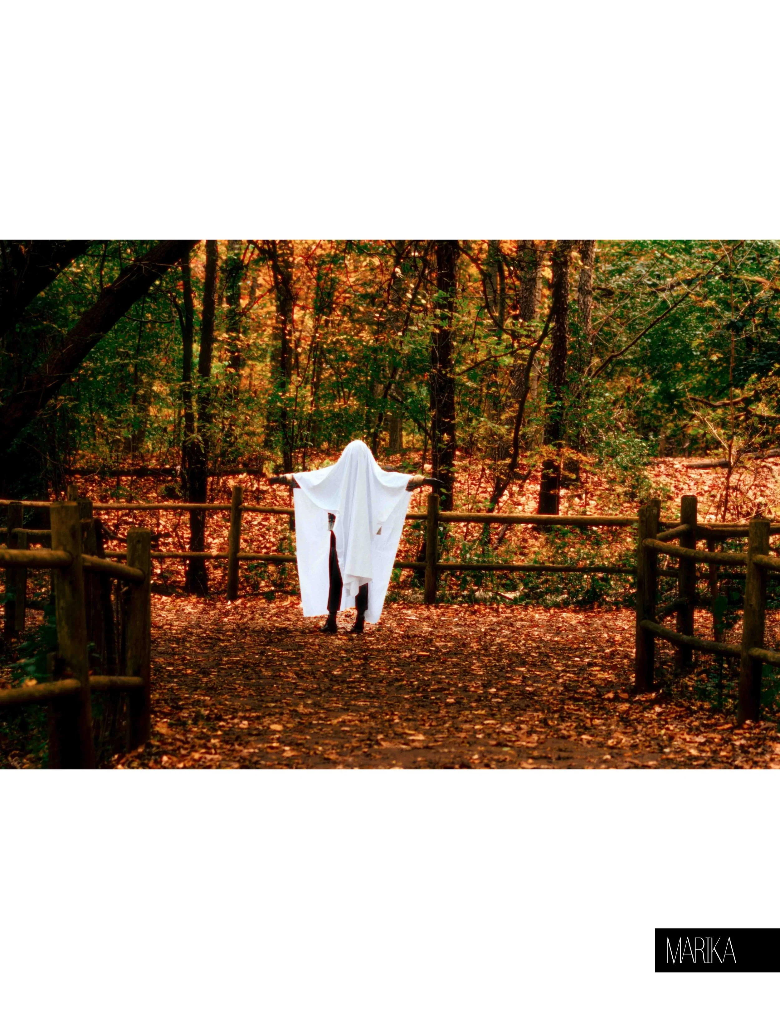 Person in a white sheet ghost costume standing on a forest trail during autumn