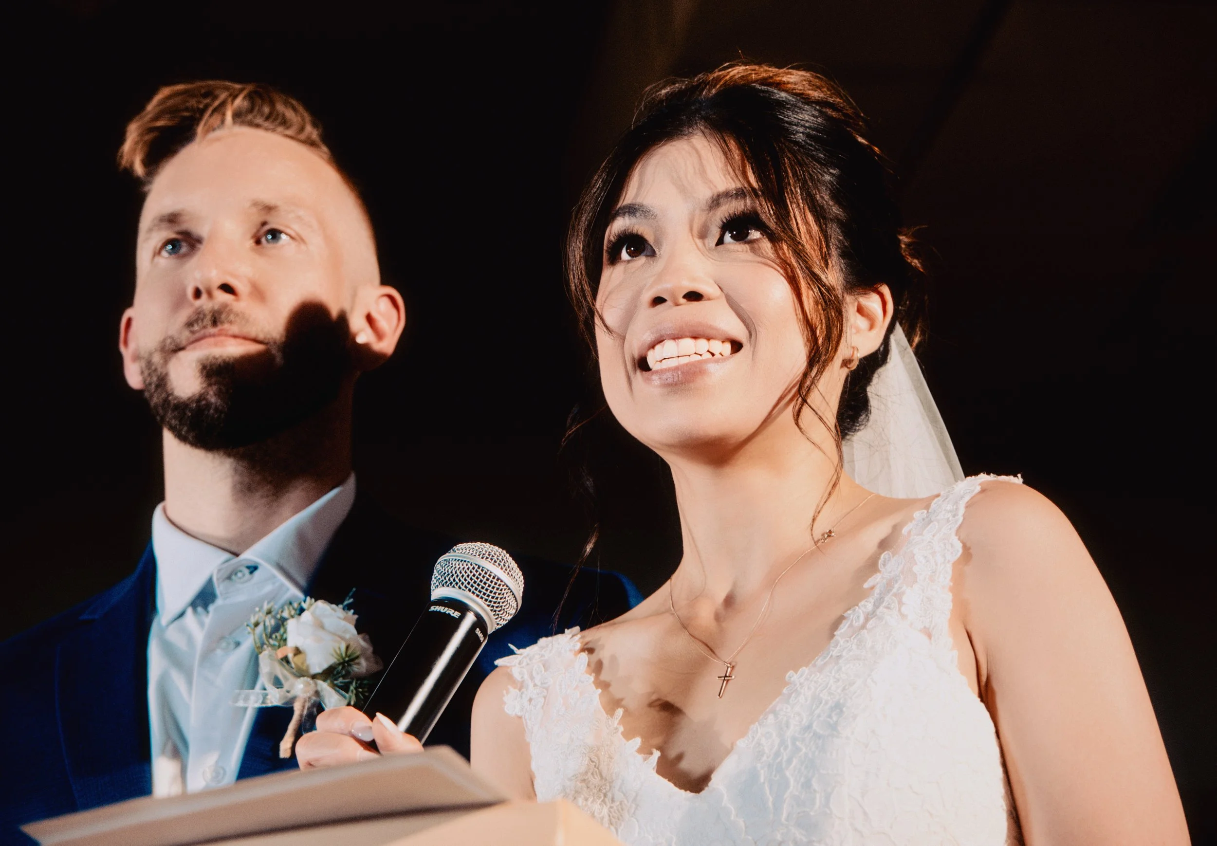 A bride and groom during their wedding ceremony. The bride, of Asian descent, is smiling with her tongue sticking out slightly, wearing a white lace wedding dress and a necklace with a cross pendant. The groom, of Caucasian descent, has a beard and m