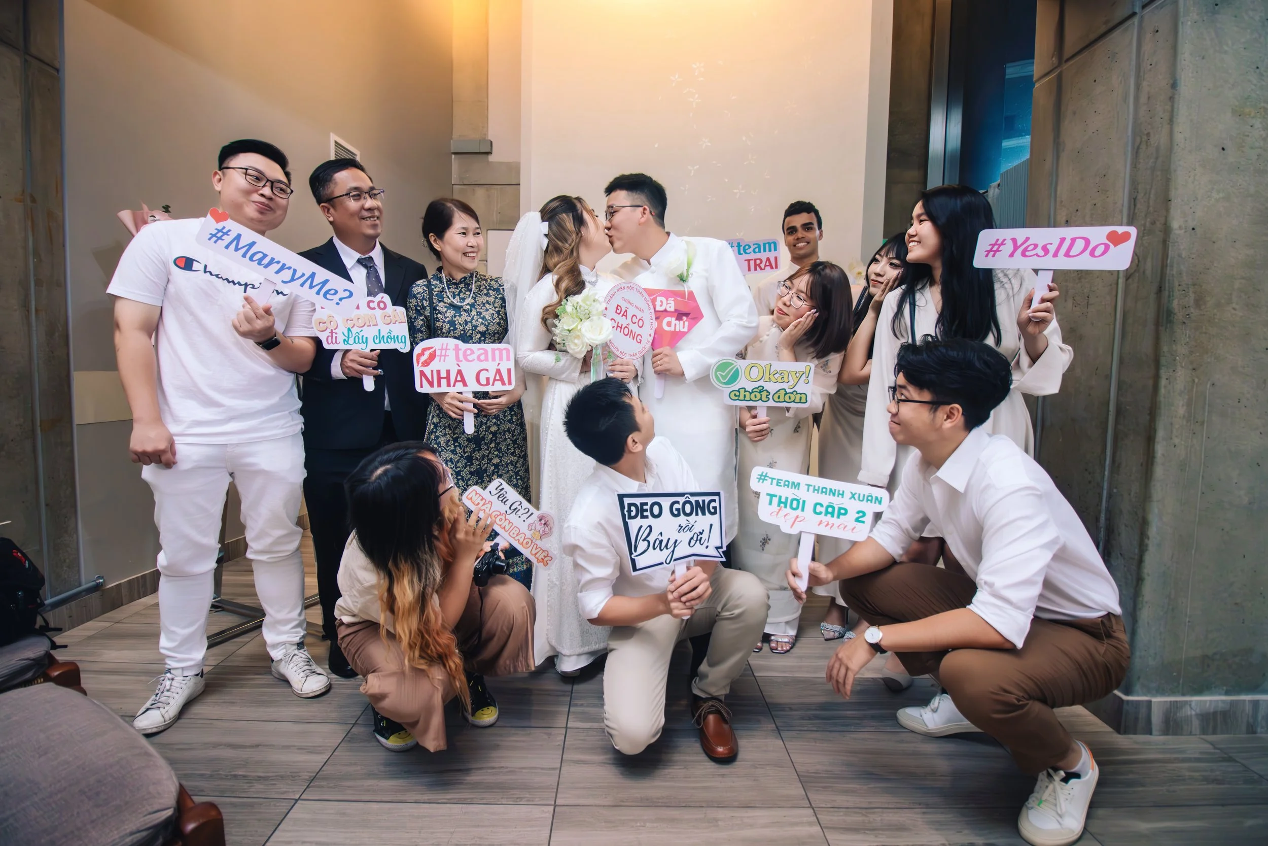 A bride and groom kissing surrounded by family and friends holding colorful signs at a wedding celebration.