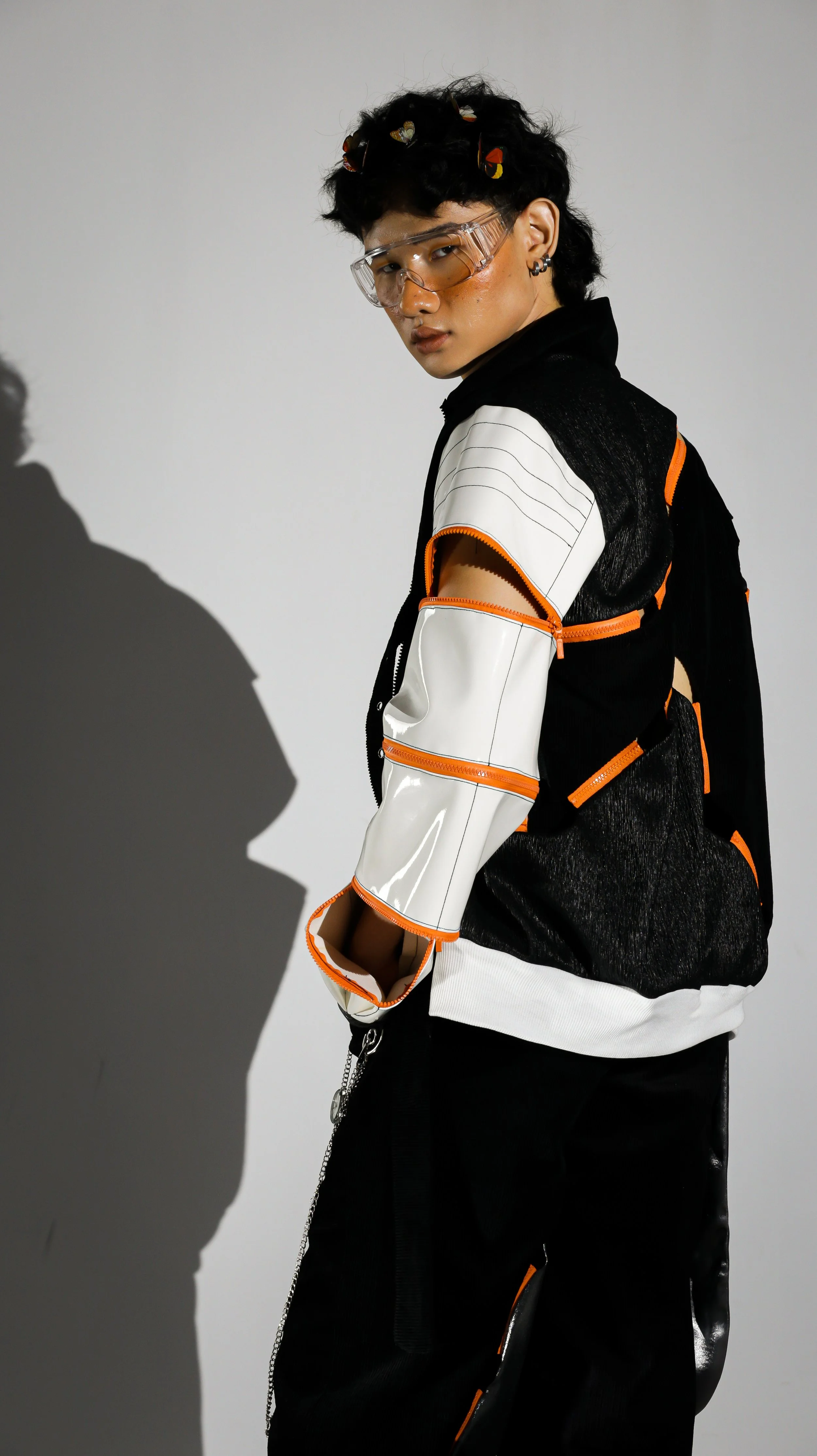 Young person with short black curly hair adorned with small butterfly accessories, wearing futuristic safety goggles, a black and white jacket with orange accents, and black pants, standing against a plain background with a shadow cast on the wall.