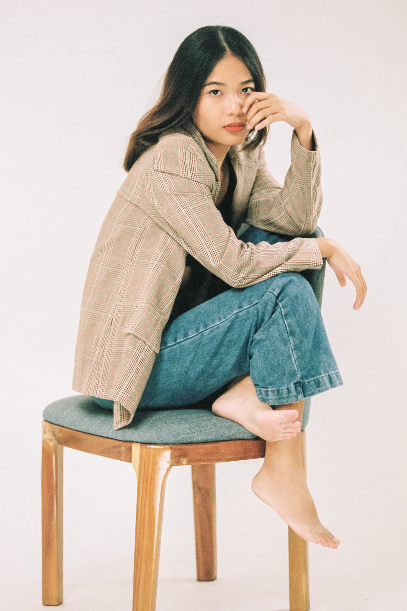 A woman sitting barefoot on a chair with one foot resting on the seat and the other hanging down, wearing a beige plaid blazer and blue jeans, with long dark hair and a serious expression.