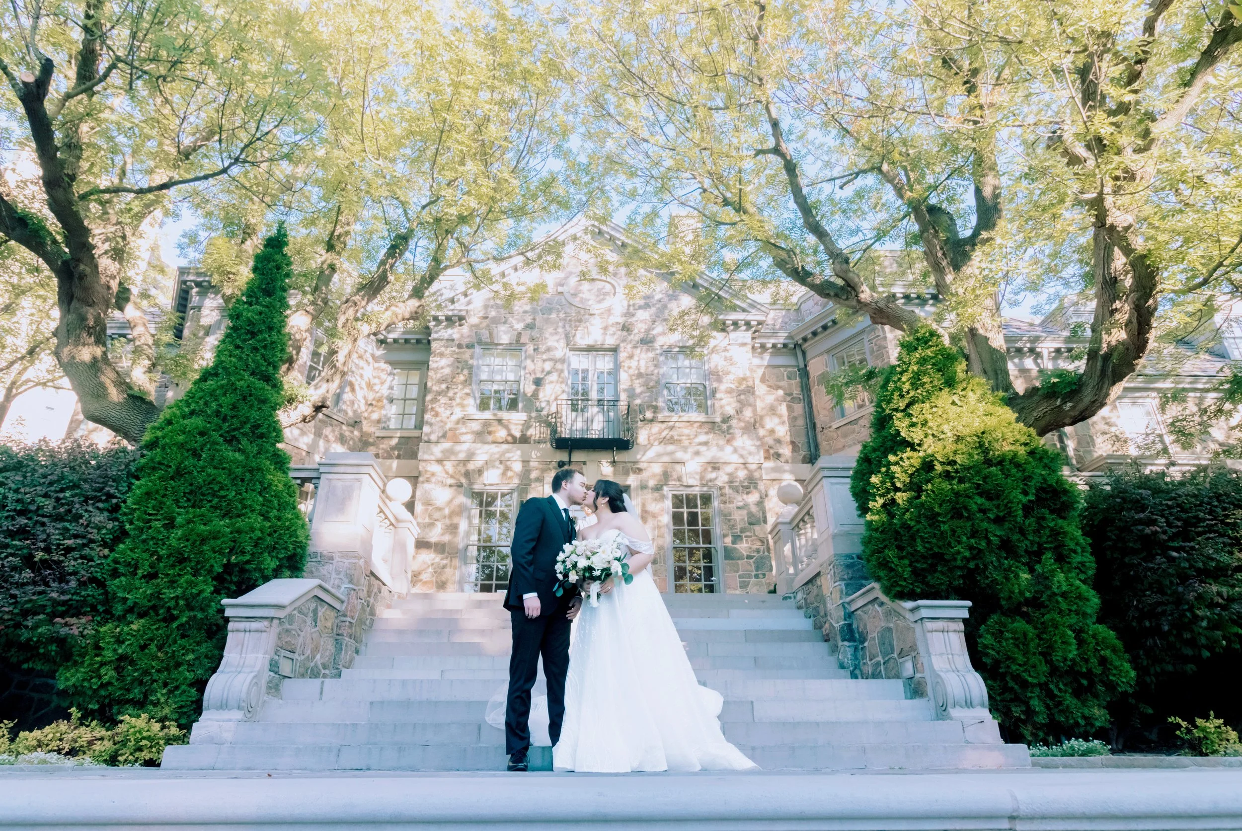 A bride and groom standing on a stone staircase outside a historic building, sharing a moment, with trees and greenery around them.