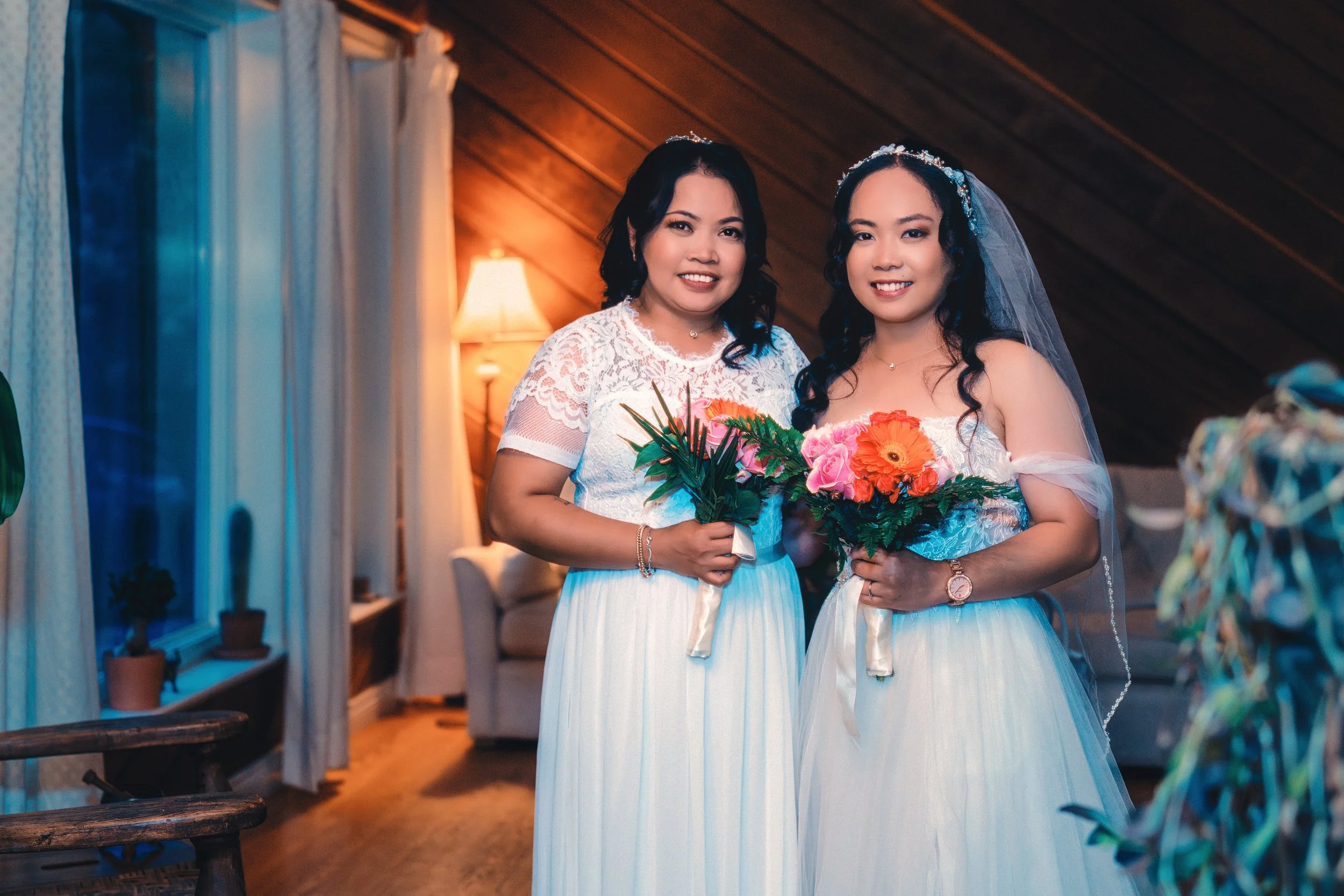 Two women in white dresses, one wearing a veil, holding colorful flower bouquets, standing in a warmly lit room with wooden walls and windows with curtains, smiling for a photo.